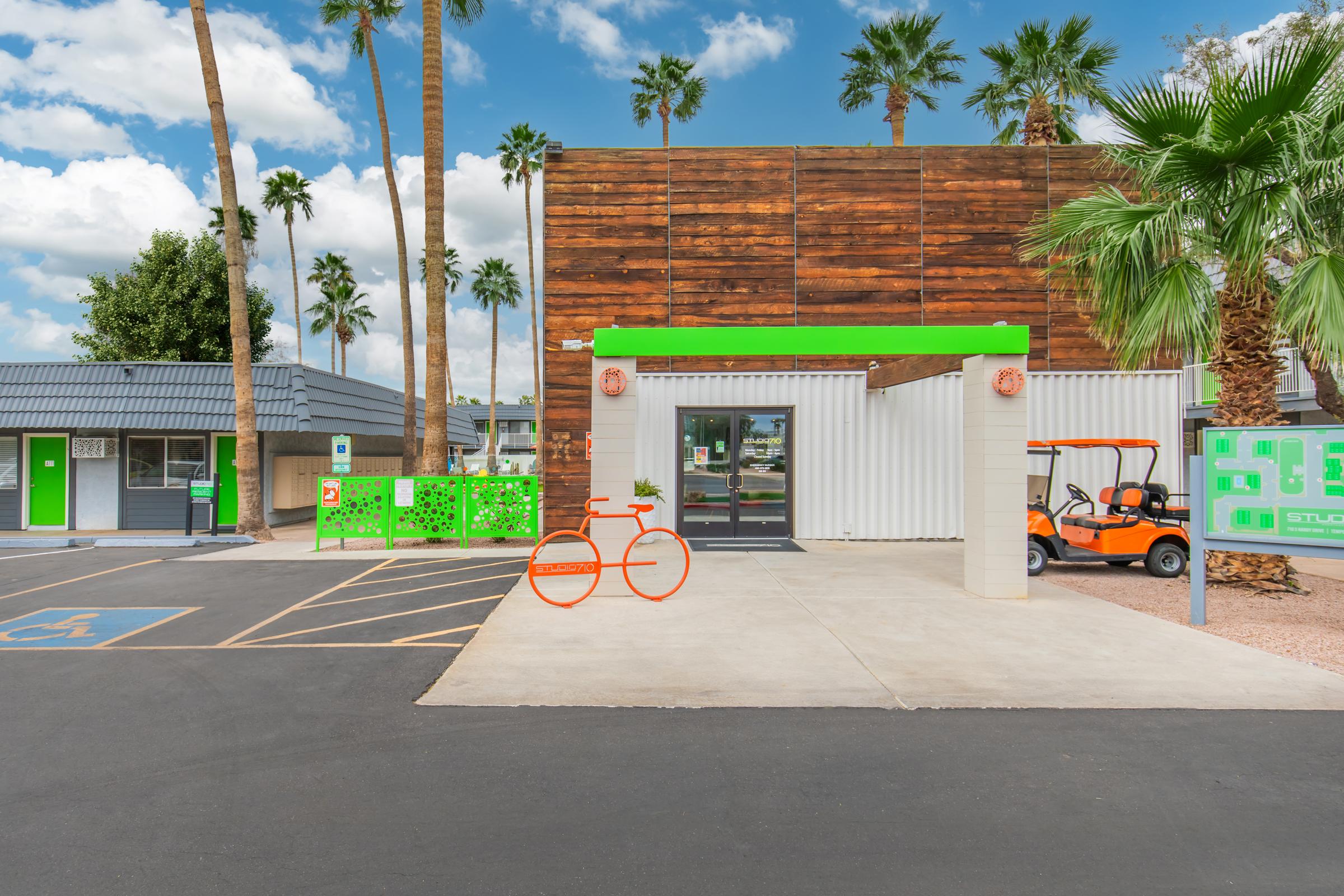 Modern storage facility entrance featuring a wooden facade and bright green accents. The area includes palm trees, parking spaces, an orange golf cart, and a decorative bicycle sculpture. The sky is blue with clouds, creating a vibrant atmosphere.
