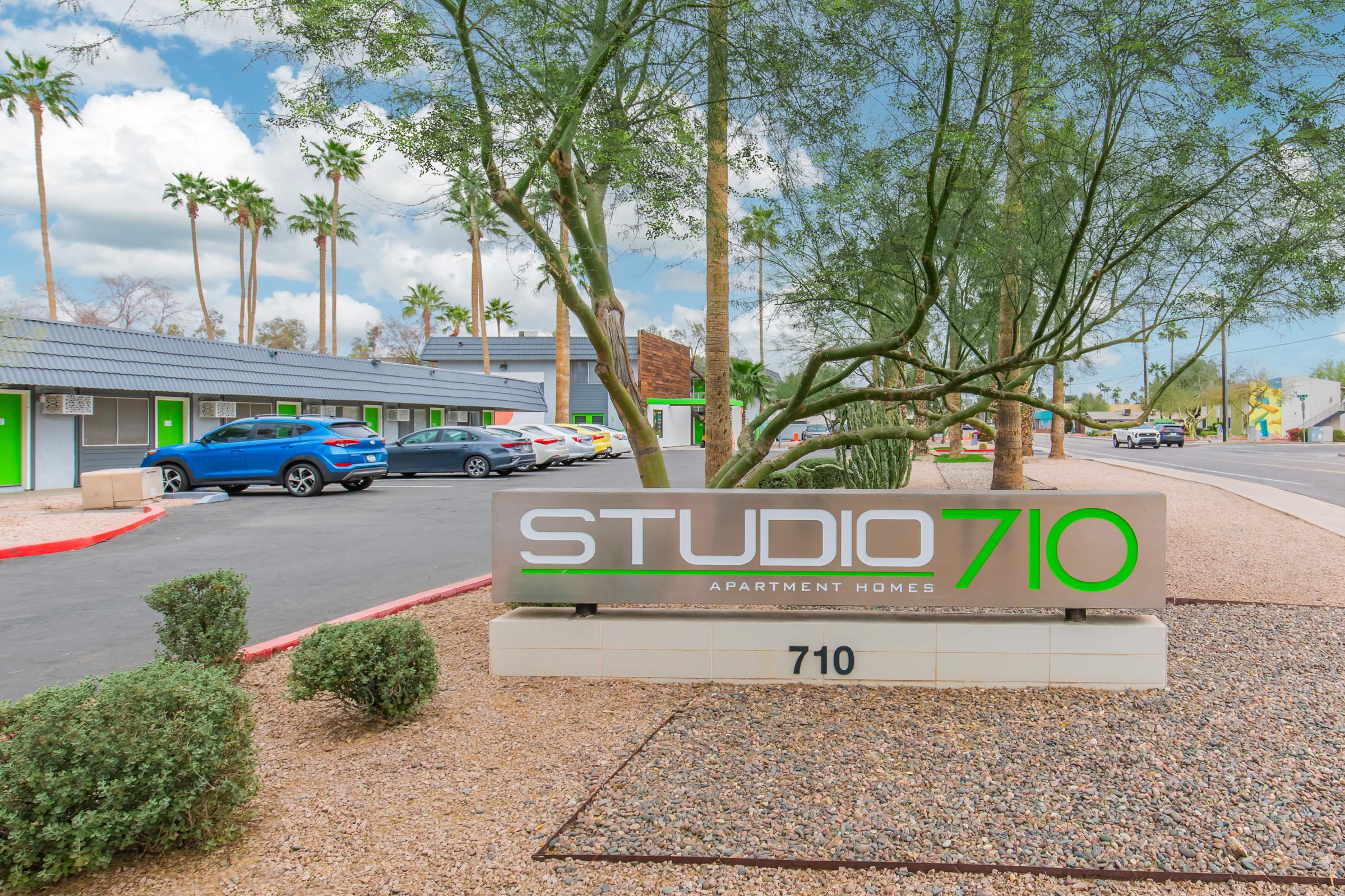 Signage for Studio 710 Apartment Homes, featuring the name "STUDIO 710" prominently displayed in green and white. Surrounding the sign are desert landscaping and palm trees, with a view of apartment buildings and parked cars in the background. The scene is bright and welcoming.