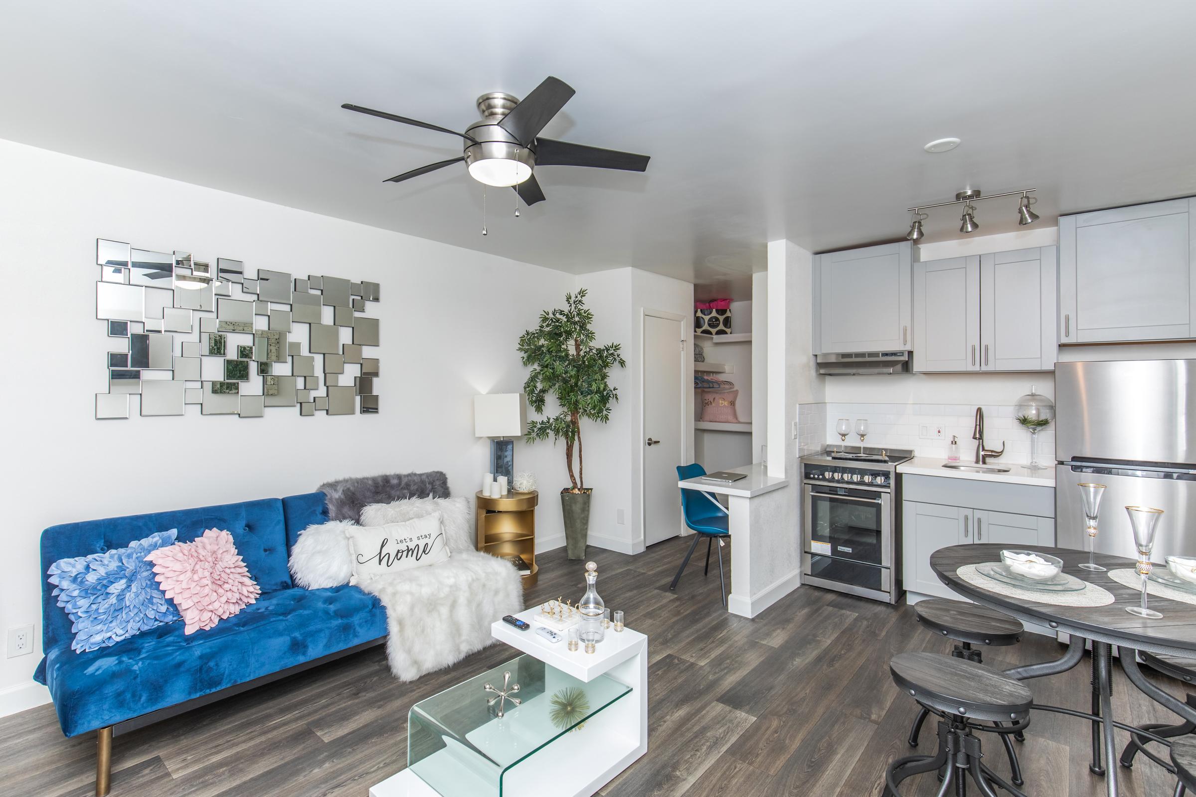 A modern living space featuring a blue velvet sofa with decorative pillows, a glass coffee table, and a small dining area with chairs. A kitchen is visible in the background with stainless steel appliances. The walls are adorned with a mirrored art piece, and a potted plant adds a touch of greenery.