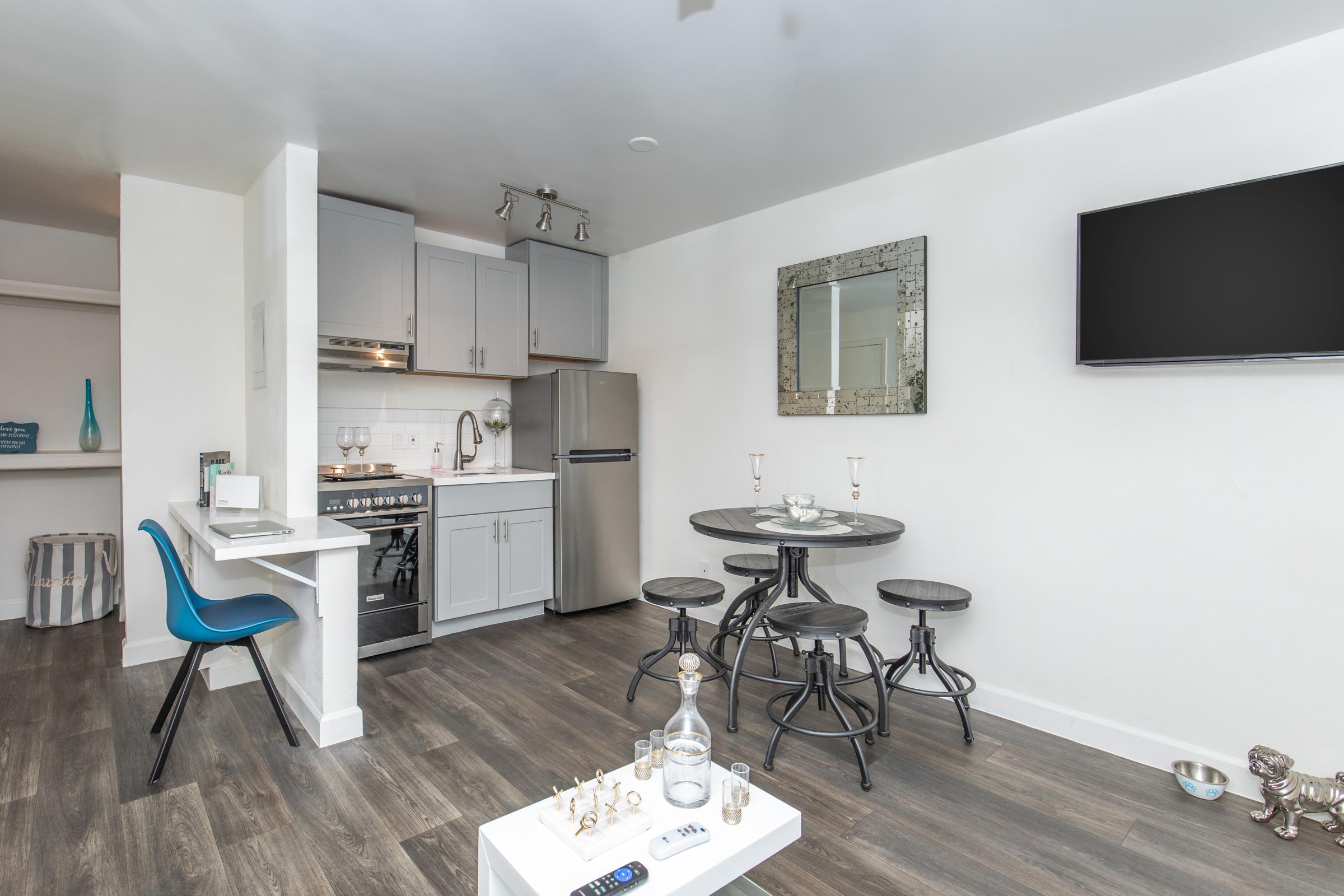 A modern kitchen and dining area featuring gray cabinets, stainless steel refrigerator, and a round dining table with black stools. The space has a minimalist design with a large mirror on the wall, a wall-mounted TV, and warm wooden flooring, creating a cozy and inviting atmosphere.
