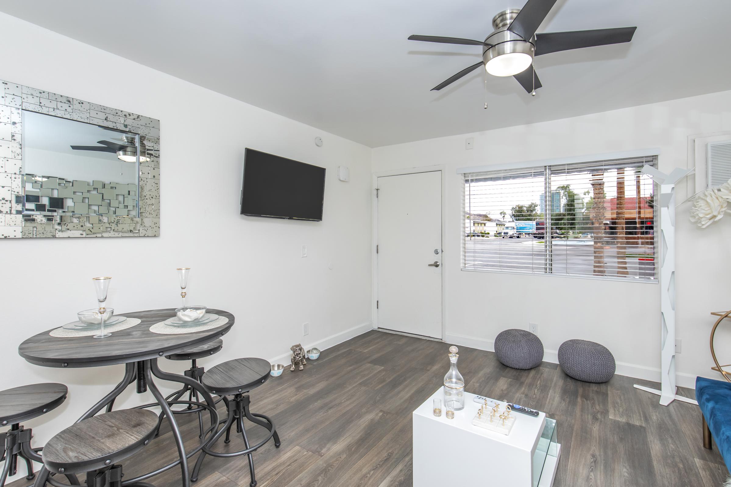 A modern living space featuring a round dining table with two place settings, a wall-mounted flat-screen TV, and a mirrored wall decor. A door leads outside, and there's a window with blinds, allowing natural light in. Two gray bean bag chairs are positioned on the floor, enhancing the cozy ambiance.