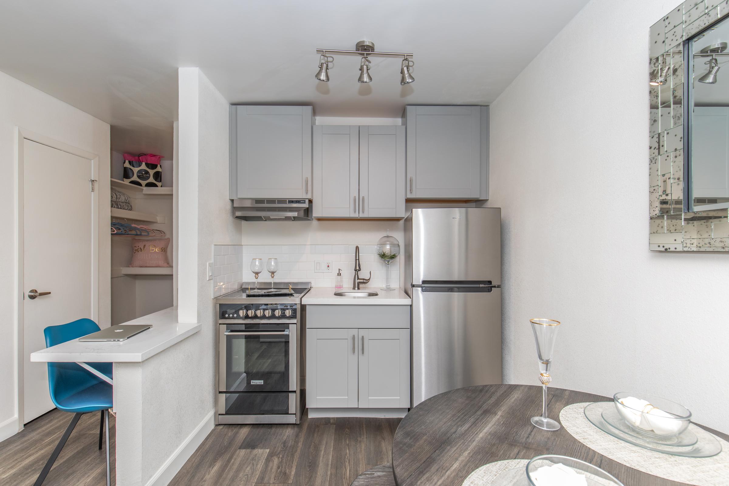 A modern kitchen featuring gray cabinets, stainless steel appliances, and a sleek refrigerator. There’s a small dining table with plates and glasses, and a minimalist design overall. The space is well-lit and includes a bar-style table with a chair in a corner.