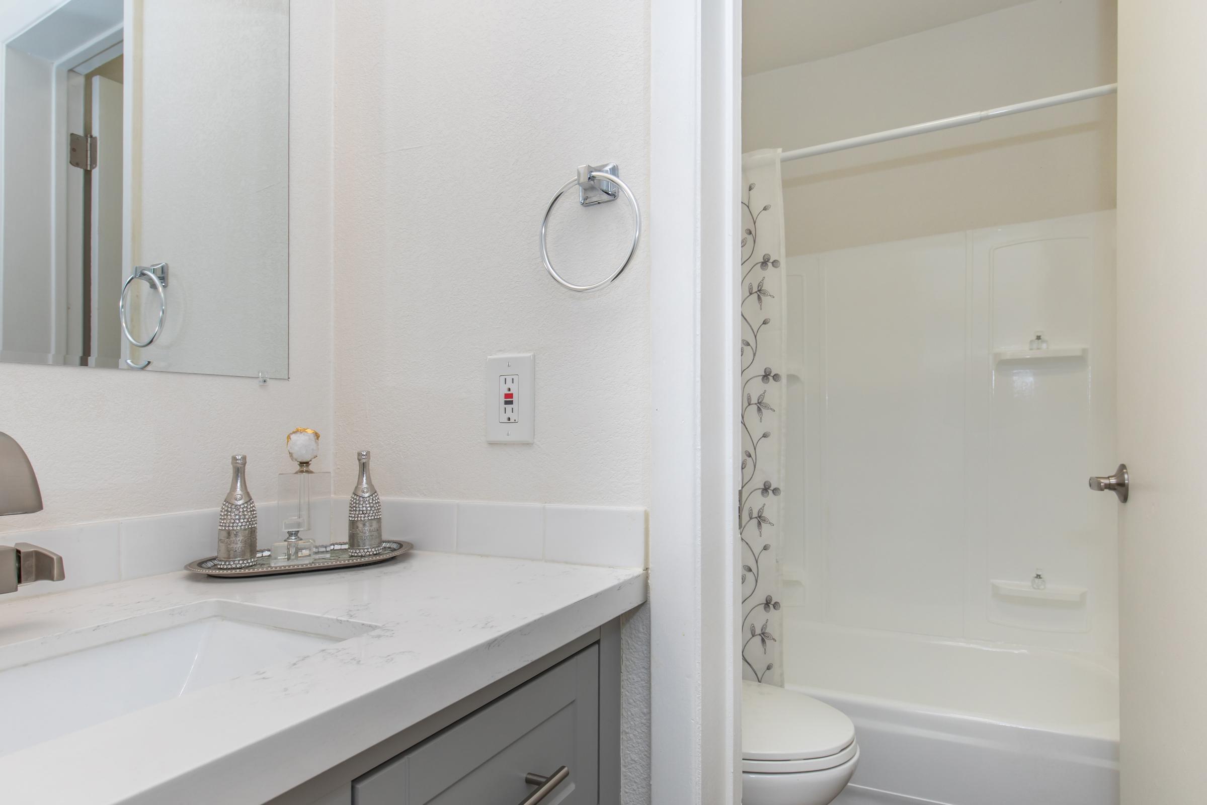 A modern bathroom featuring a sink with gray cabinetry, a mirror above, and a towel ring installed on the wall. A shower area with a white tub and simple patterned shower curtain is visible to the right. The overall color scheme is light and neutral, creating a clean and airy atmosphere.