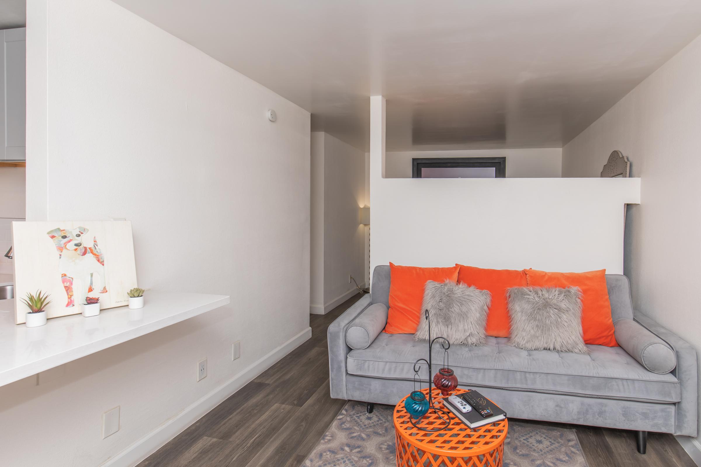 A modern living space featuring a light gray sofa with orange accent pillows and decorative throw pillows. There is a round orange coffee table with a magazine on it. The walls are white, and a small table with potted plants is visible in the background, along with a glimpse of a kitchen area.