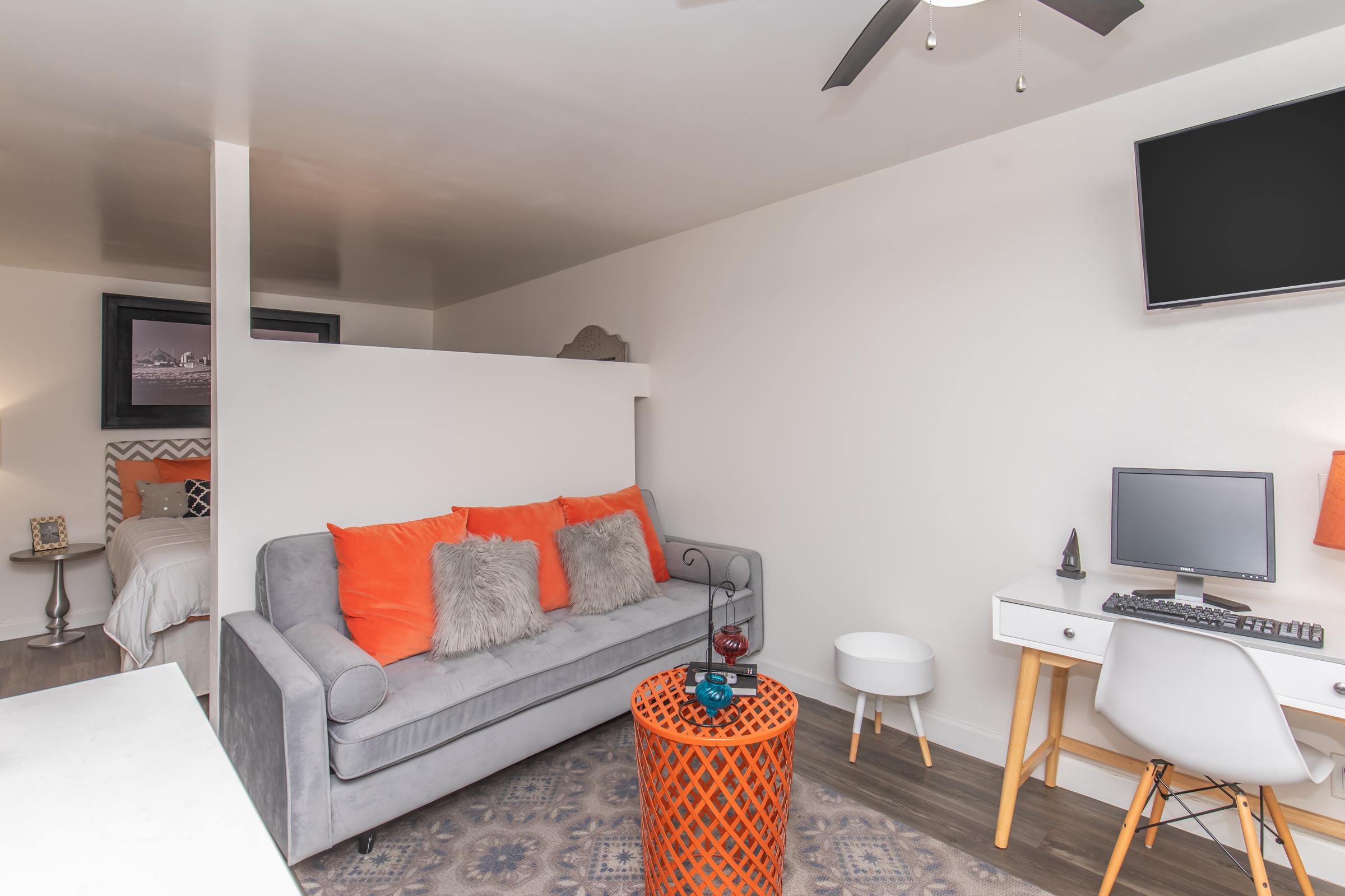 A cozy, modern living space featuring a gray sofa with orange throw pillows, a decorative circular table, a small white desk with a computer, and a wall-mounted TV. The room has a light color scheme and includes a bedroom area in the background, visible through an open space.