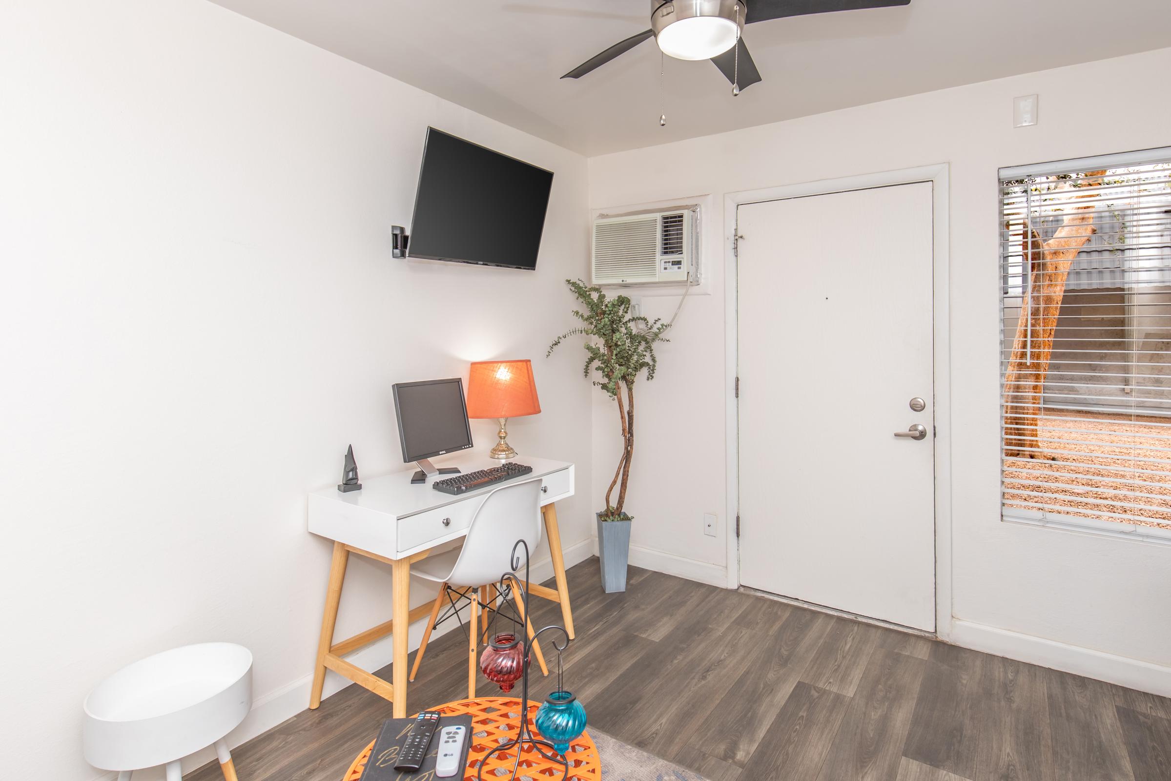 A small, modern room featuring a white wall, light wood flooring, and a ceiling fan. There's a mounted television, an air conditioning unit, a desk with a computer, a lamp on the desk, and a small round table. A window with blinds provides a view of an outdoor area with a tree.
