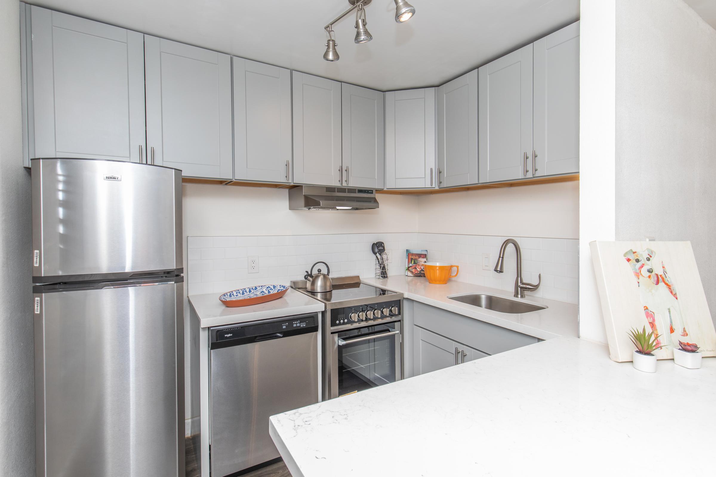 Modern kitchen with gray cabinets, stainless steel appliances including a fridge, dishwasher, and oven. A white countertop with a sink is visible, along with a kettle and an orange bowl. There's a decorative piece on the counter and a colorful artwork on the wall. Bright and inviting atmosphere.