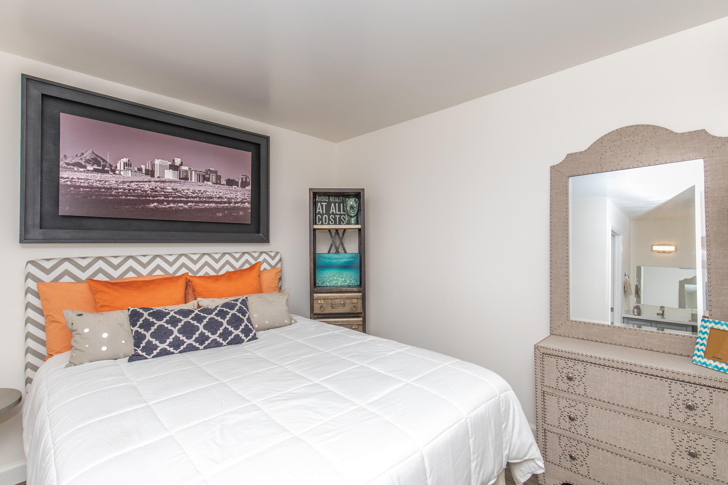 Cozy bedroom featuring a queen-sized bed with white bedding and a variety of decorative pillows. A large framed black-and-white city skyline photograph hangs on the wall above the bed. To the side, there is a stylish dresser with a mirror and decorative accents, creating an inviting atmosphere.