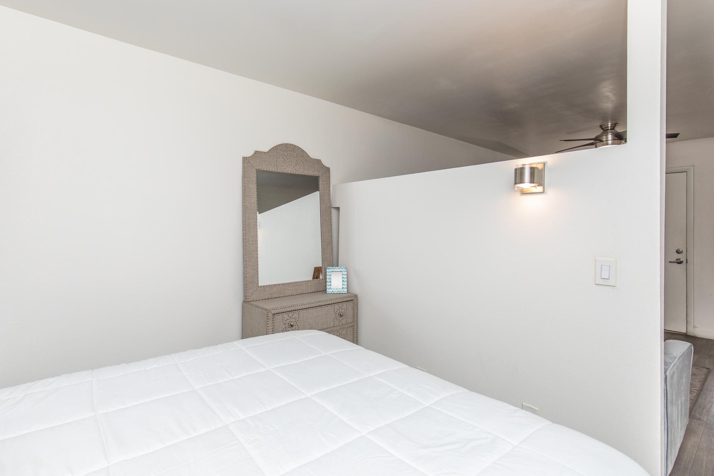 A bright and minimalistic bedroom interior featuring a neatly made bed with a white comforter. A decorative mirror is positioned on a dresser against a light-colored wall, and soft lighting from a wall fixture adds warmth to the space. A small side table displays a digital clock or device. The room has a modern, clean aesthetic.