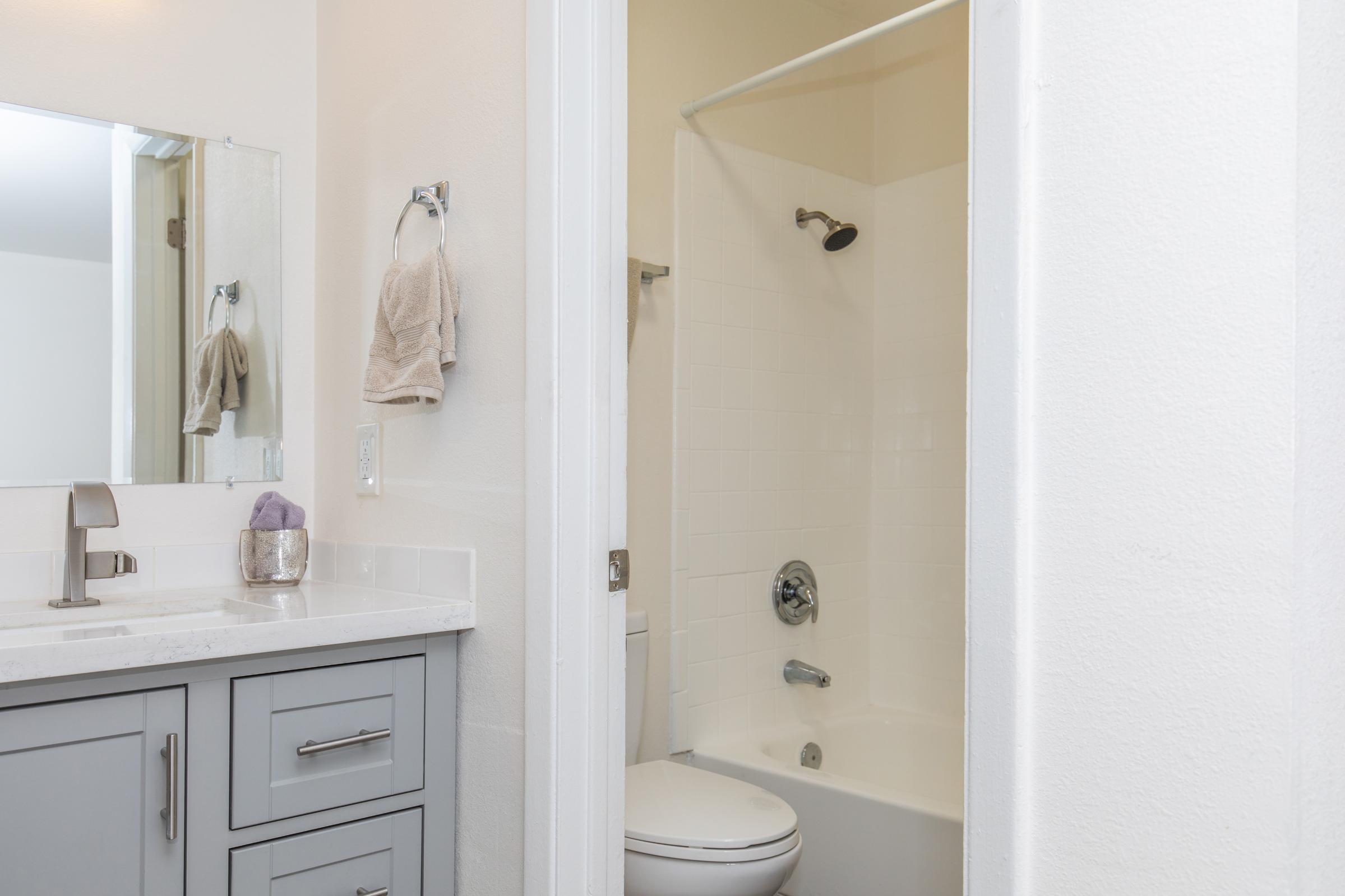A modern bathroom featuring a shower and bathtub combo, a toilet, and a vanity with a sink. The vanity has a mirror above it, and a hand towel is hanging nearby. The color scheme is light and neutral, creating a clean and spacious appearance.