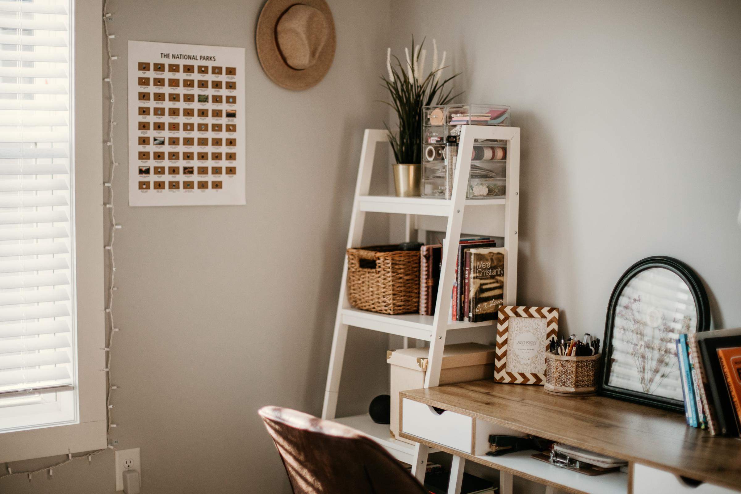 A cozy workspace featuring a wooden desk with stationery, a round mirror, and books. Behind, a white shelf holds decorative baskets and books, alongside a framed poster titled "The National Parks." Natural light filters through a window adorned with blinds, and a hat hangs on the wall.