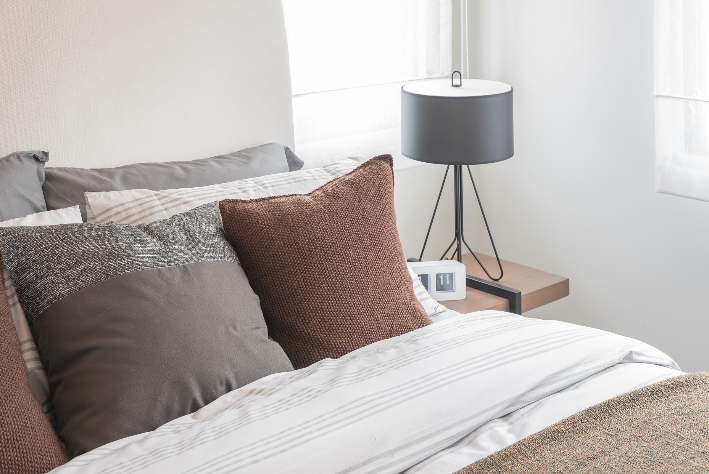 A neatly made bed featuring gray and brown pillows, with a bedside table holding a modern lamp and an alarm clock. Natural light streams in through sheer curtains, creating a cozy and inviting atmosphere in the bedroom.