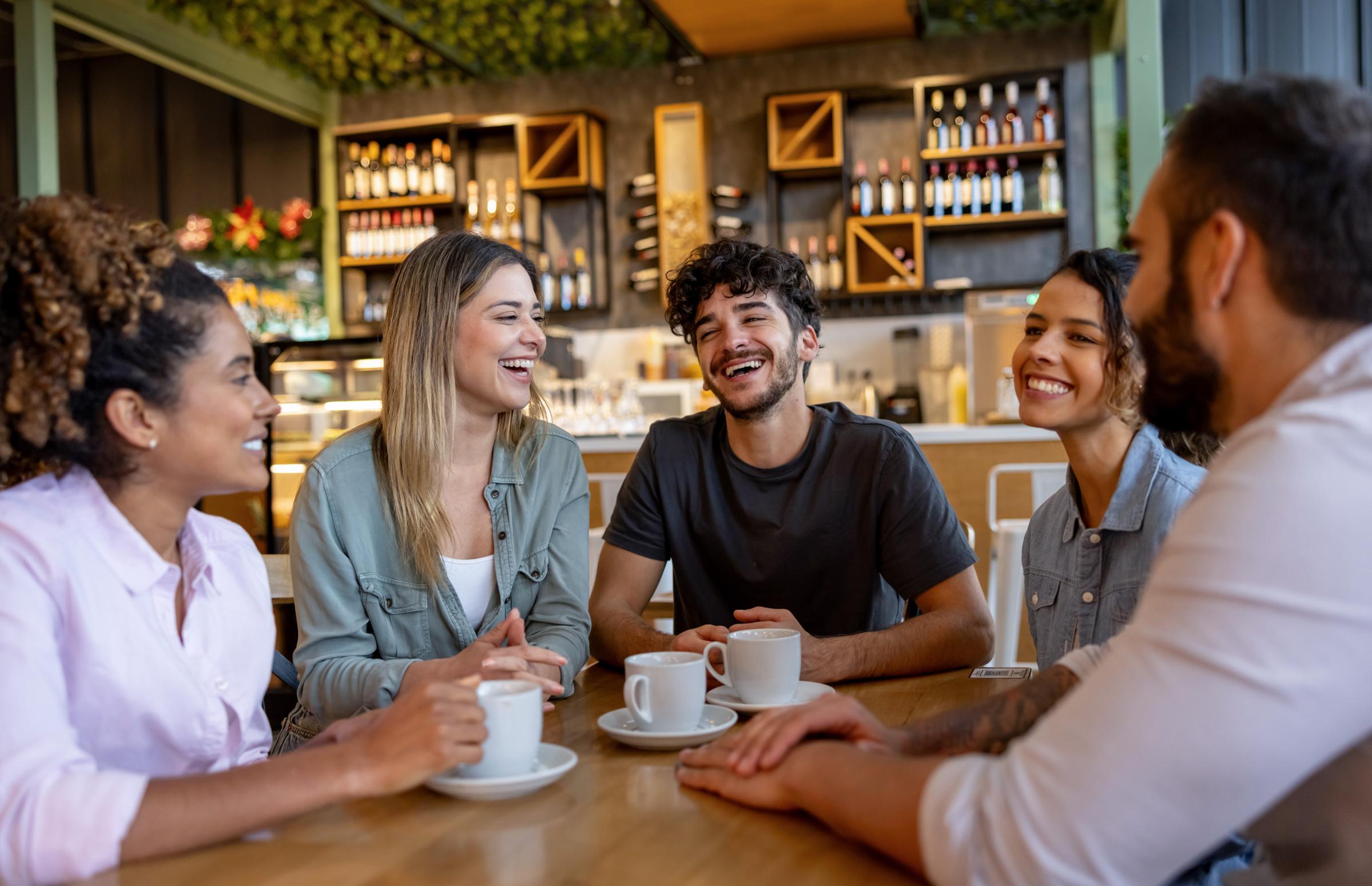 A group of five diverse friends sitting around a wooden table in a cozy café, smiling and engaging in conversation. They each have a coffee cup in front of them, creating a warm and friendly atmosphere. The café interior features shelves with bottles and plants, enhancing the inviting vibe.