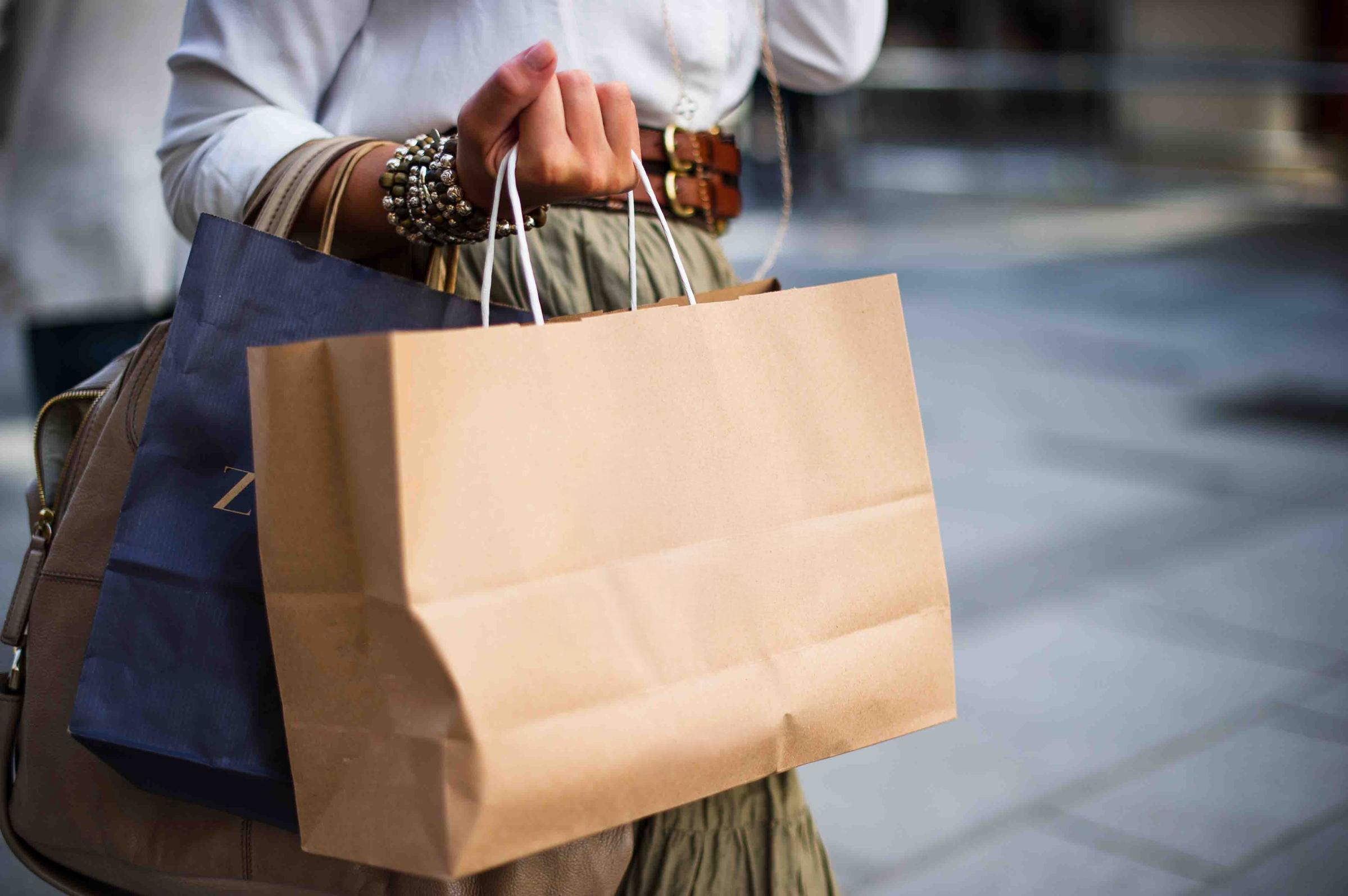 A close-up of a person’s hand holding a brown paper shopping bag, with a blue bag partially visible in the background. The individual is dressed in a white shirt and a long skirt, accessorized with bracelets. The scene is set in an urban environment.