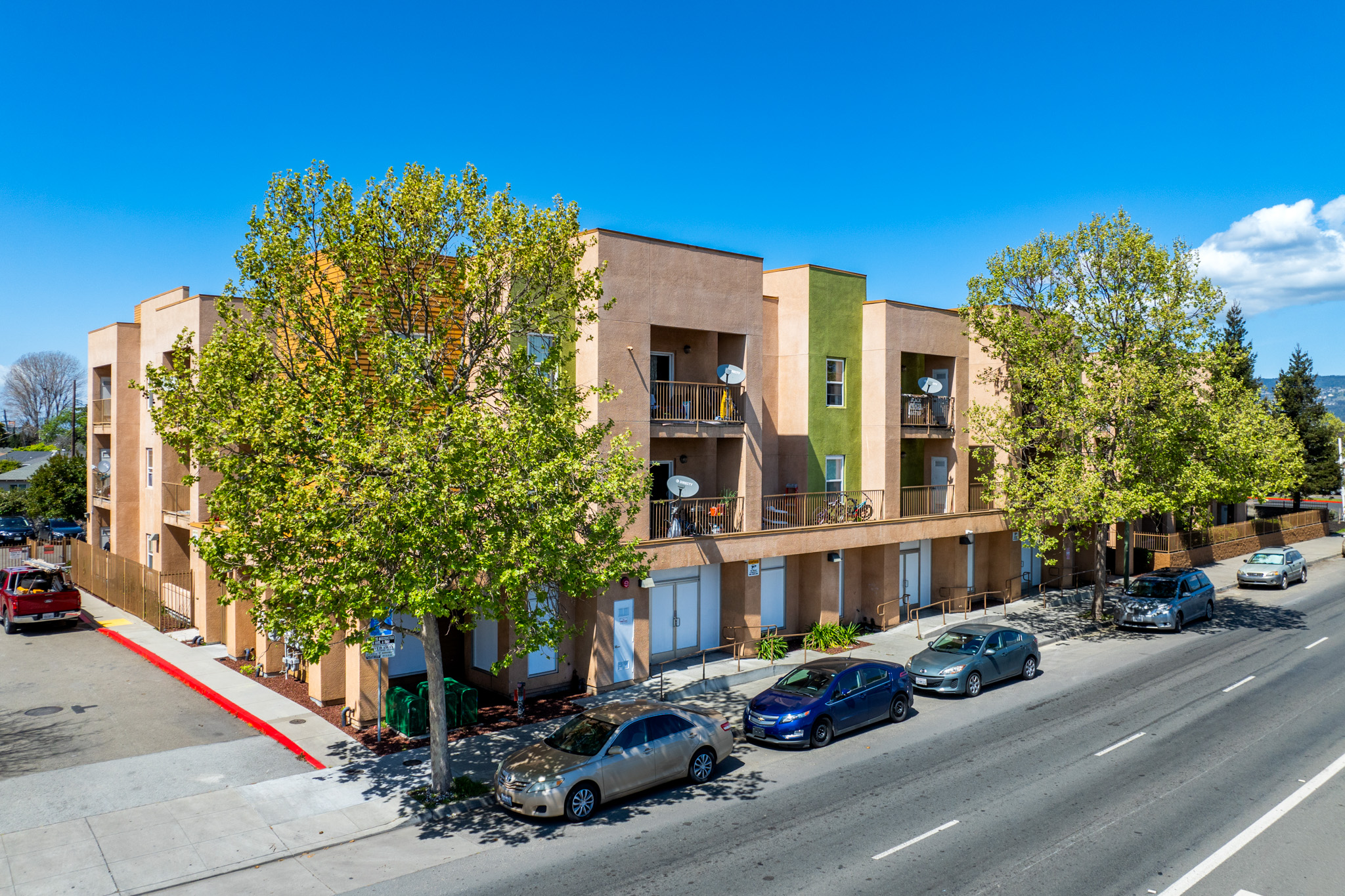 A view of a residential apartment building with multiple stories, featuring balconies and green trees along the sidewalk. Several parked cars are visible on the road. Clear blue sky with a few clouds in the background.