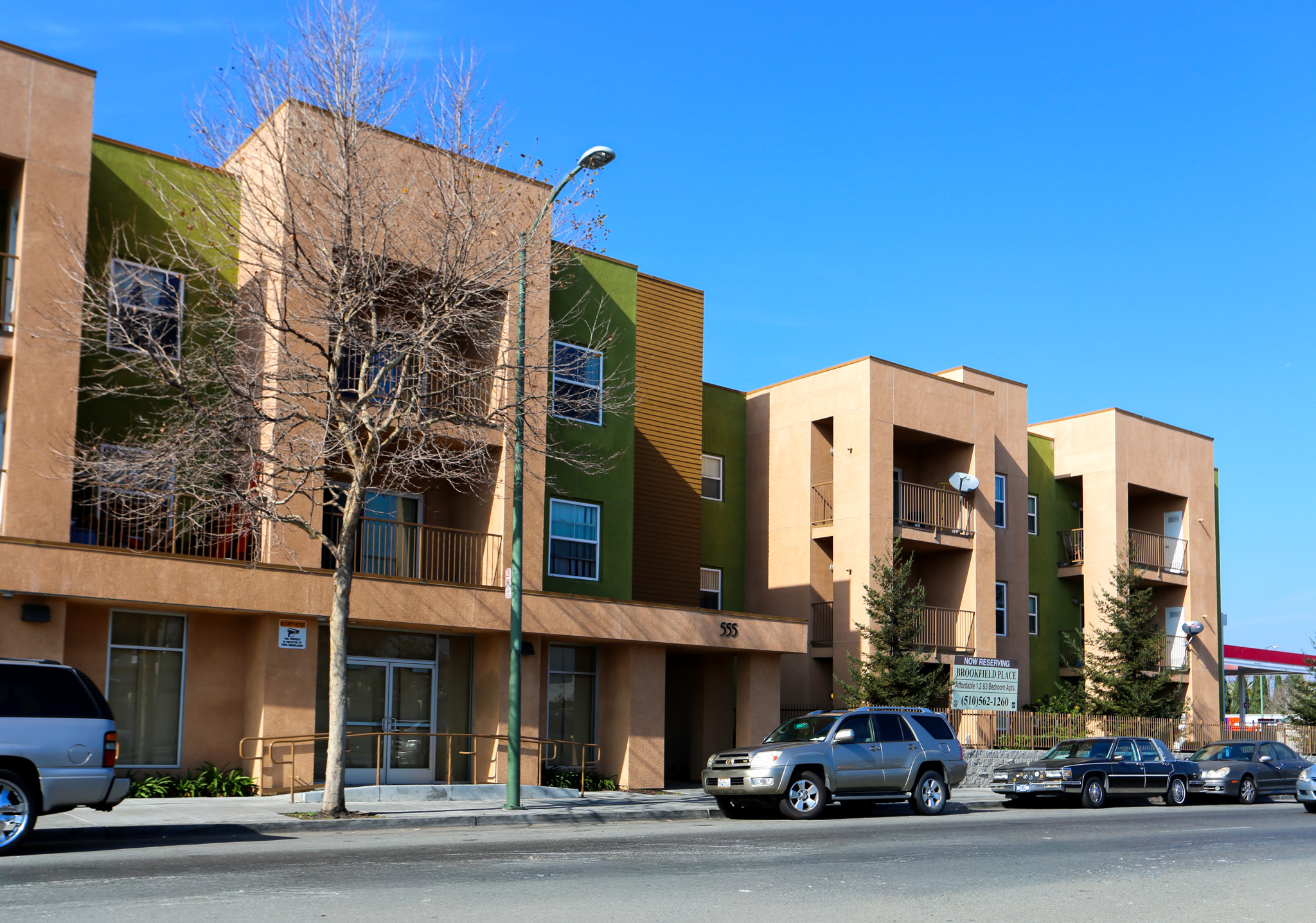 A street view of a modern apartment complex featuring a mix of beige and green exterior walls. The building has multiple windows and balconies. Parked cars are lined up along the street, and there are trees without leaves in front of the building under a clear blue sky.