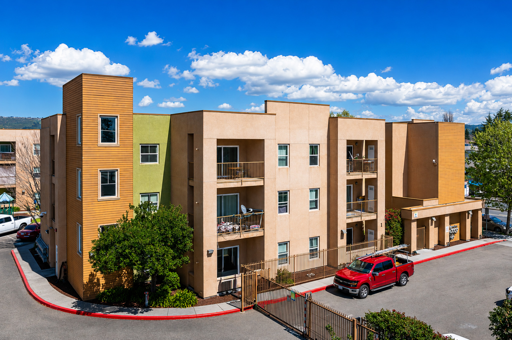 A three-story apartment building featuring a mix of beige and green façades. The building has balconies on several units, with a red truck parked in front and a gated entrance. The scene is set against a bright blue sky with fluffy white clouds and greenery surrounding the area.