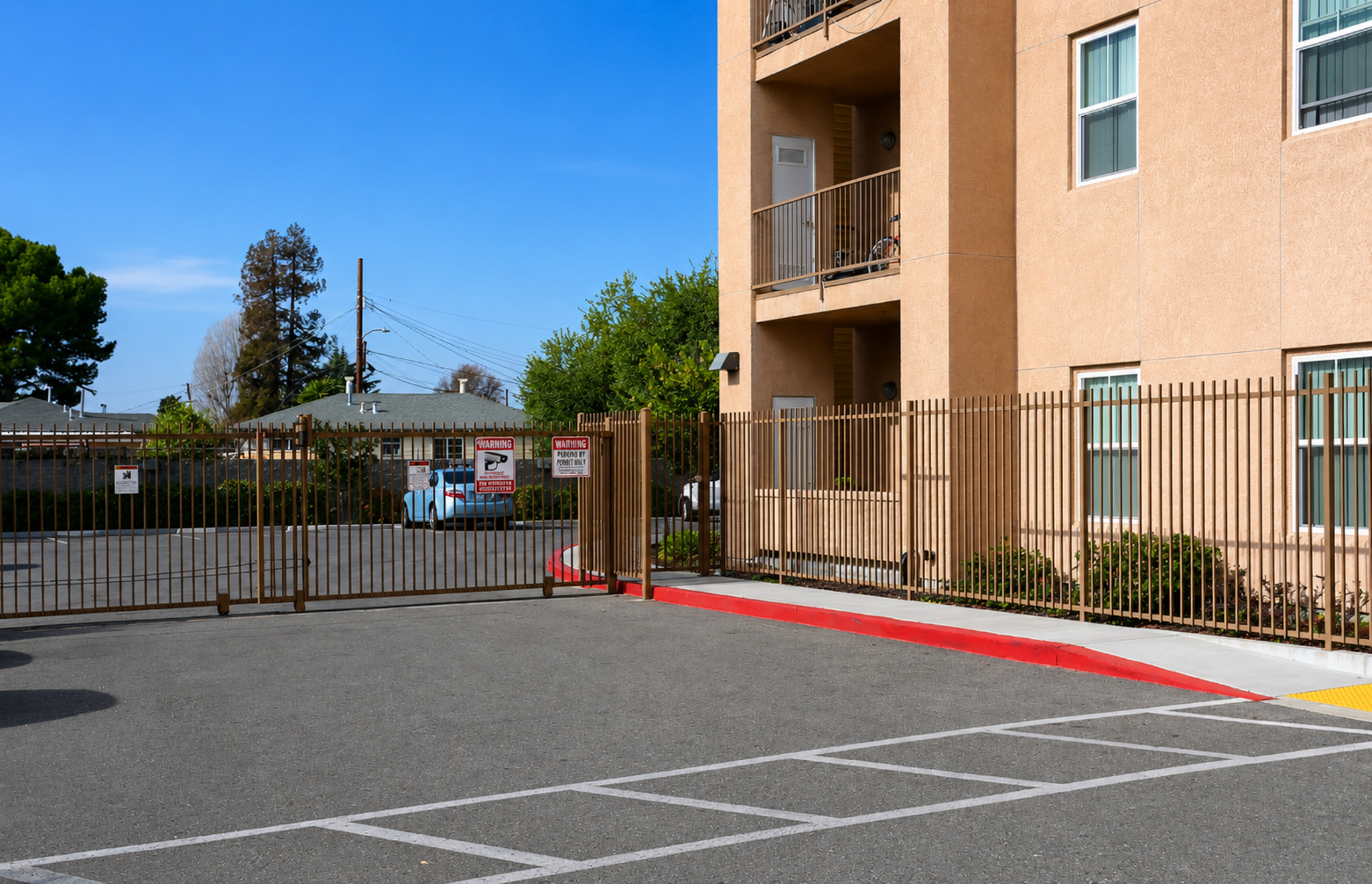 A gated entrance to a parking lot adjacent to a multi-story building. The lot features marked parking spaces, a red curb, and a gravel area in the background. There are trees and other buildings visible in the distance under a clear blue sky.