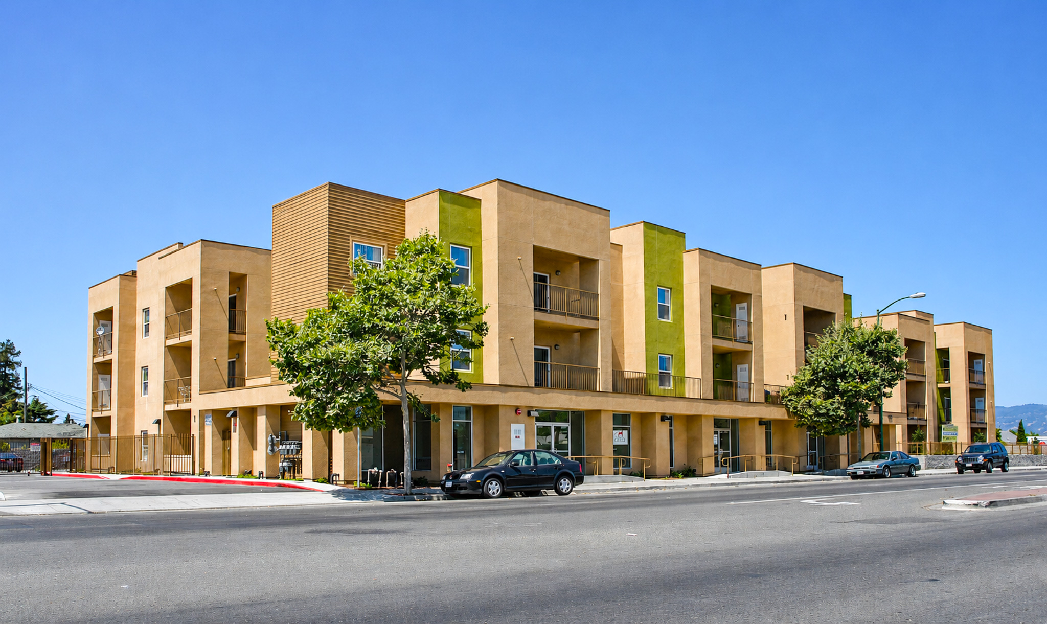 A modern multi-story building with a tan exterior and green accents, located at a street corner. It features balconies on the upper floors, large windows, and is surrounded by trees. There are parked cars on the street and a clear blue sky above. The setting appears urban and well-maintained.
