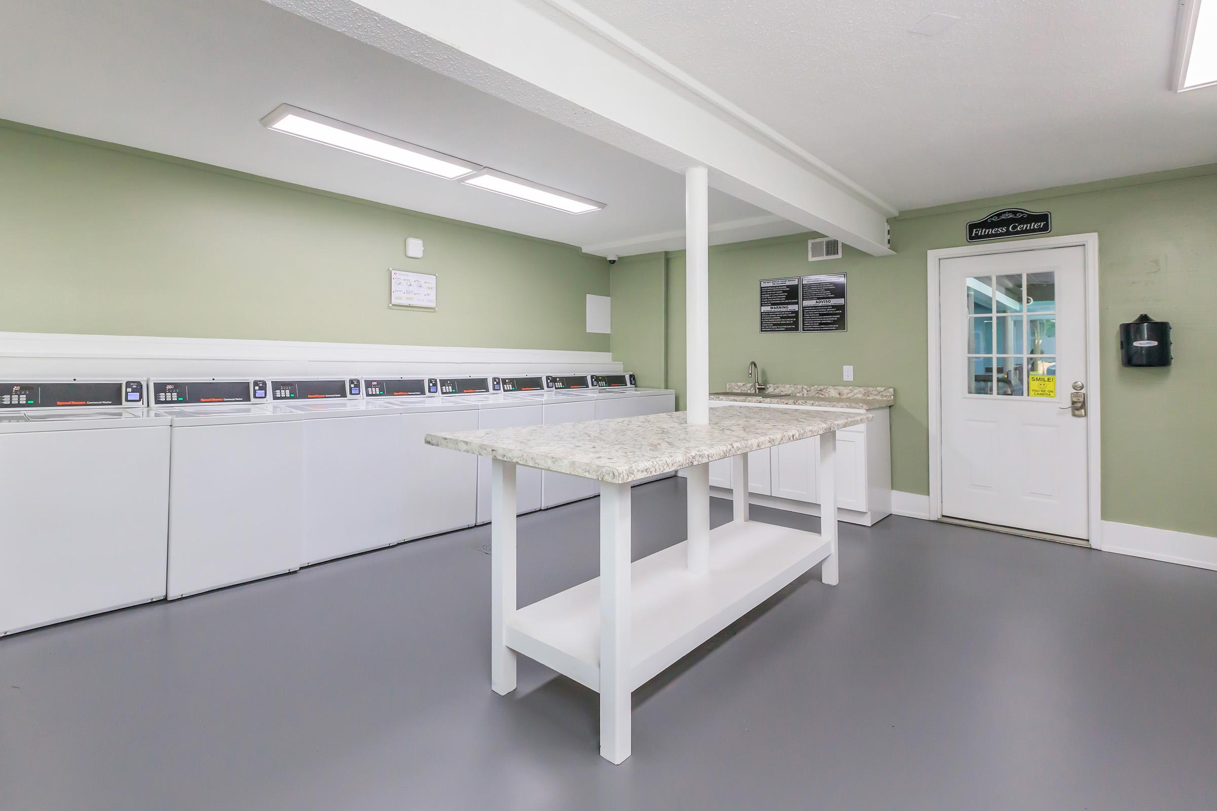 A clean and modern laundry room featuring several white washing machines, a spacious countertop in the center, and a door leading to the outside. The walls are painted in a soft green, and there are bright overhead lights enhancing the overall bright and inviting atmosphere.