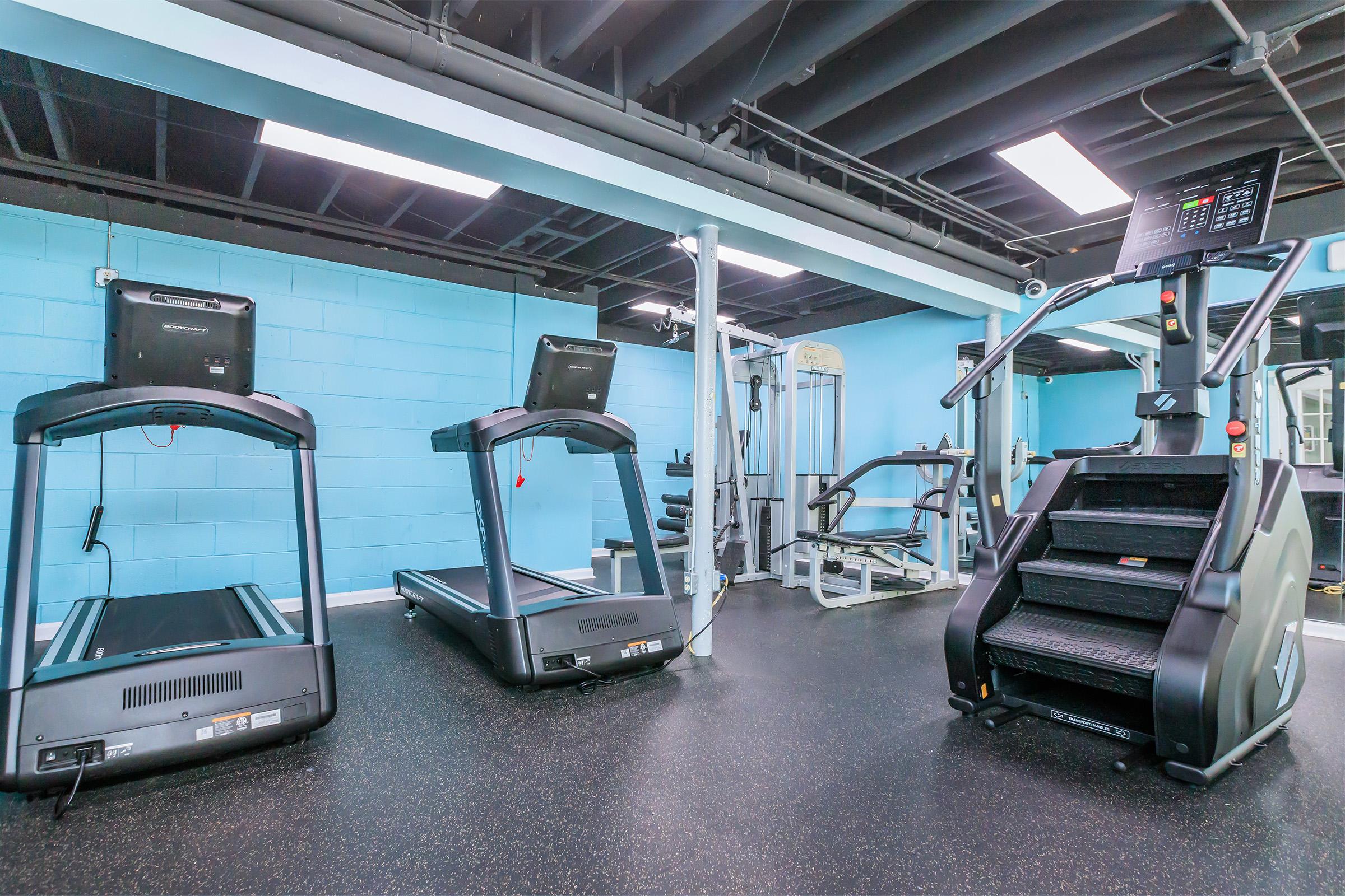 A well-lit gym interior featuring three treadmills, a stair climber, and various weightlifting equipment. The walls are painted light blue, and the flooring is dark rubber, providing a modern and inviting workout space.