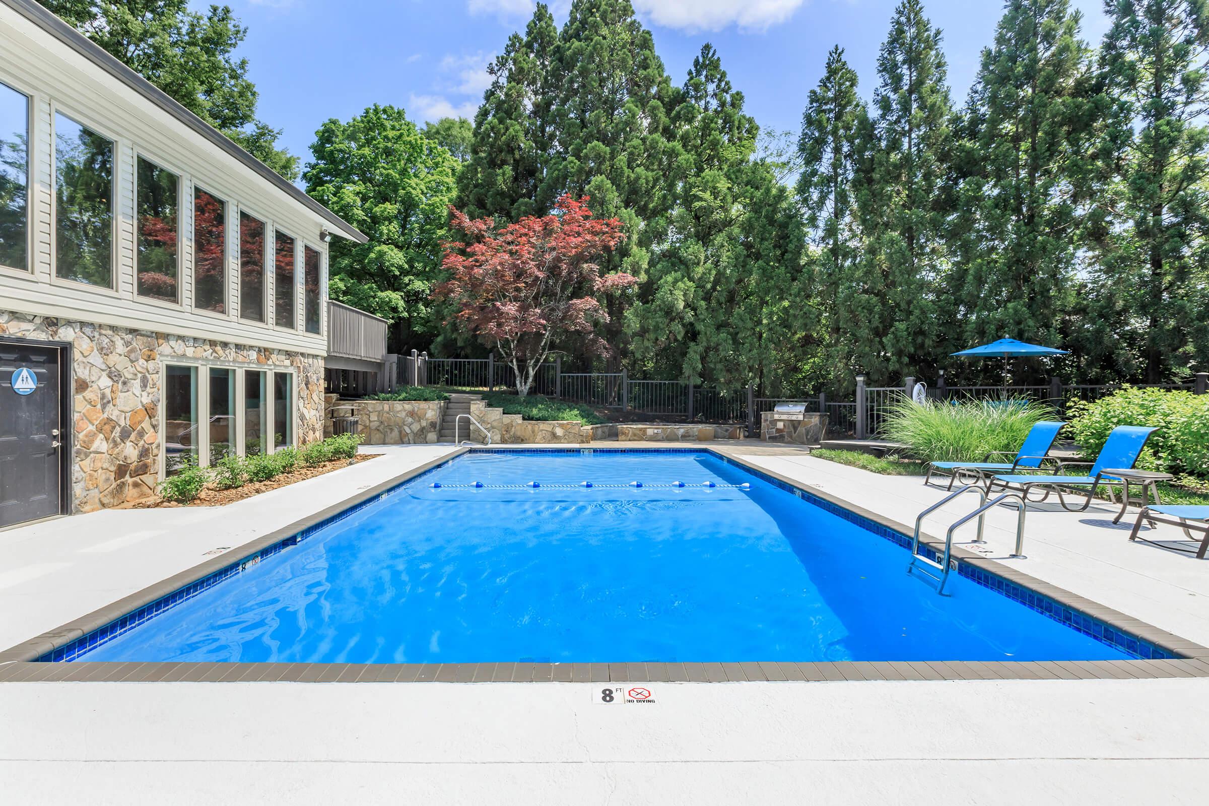 A bright, sunny swimming pool surrounded by a paved patio. The pool features a metal ladder and six blue lounge chairs along the edge. Lush green trees and shrubs provide privacy around the space, with a small poolside area that includes a shaded umbrella. A modern building with large windows is visible nearby.
