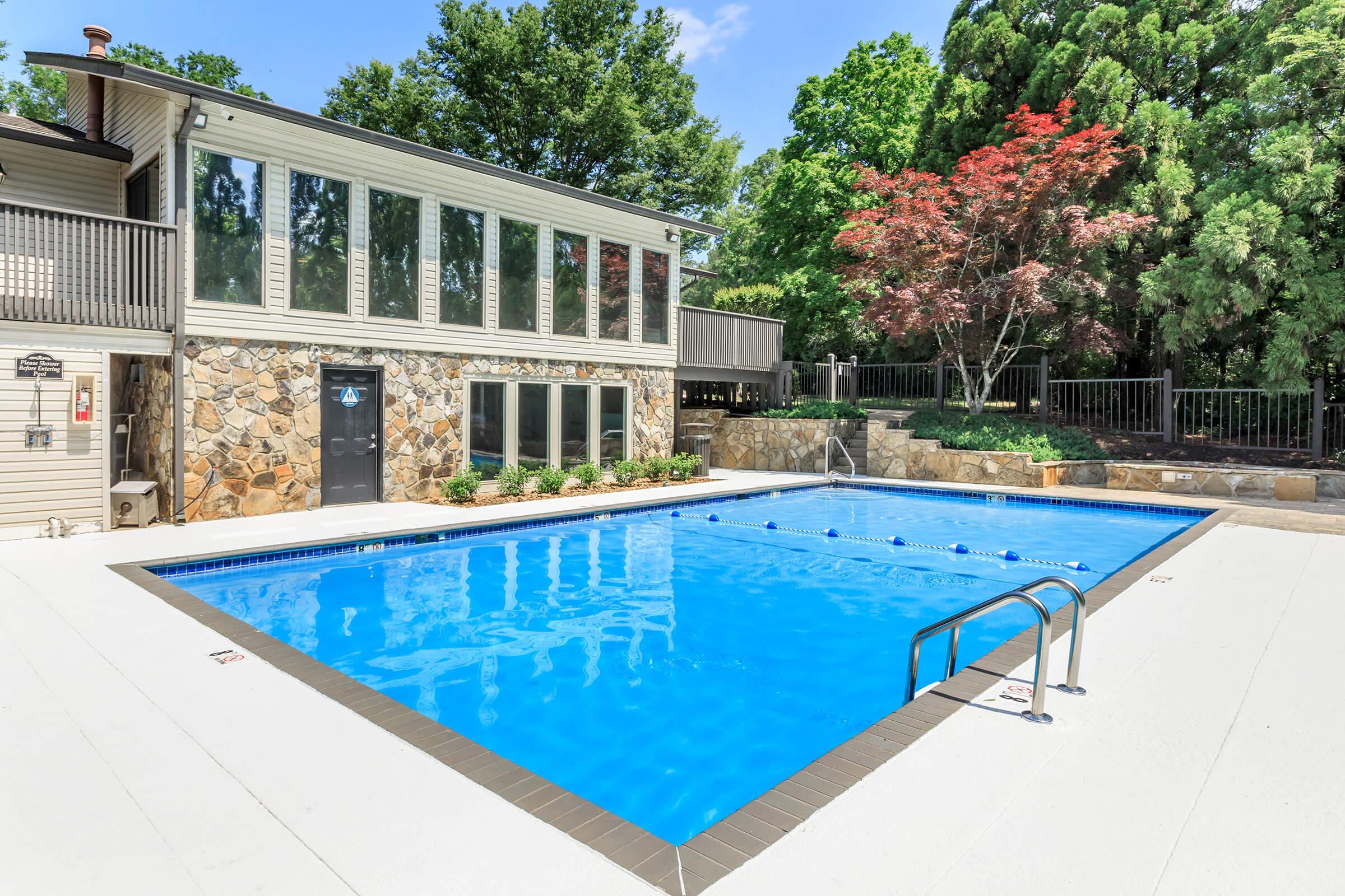 A clear blue swimming pool surrounded by a clean white deck, with a stone wall on one side and a modern building featuring large windows in the background. Lush green trees and a colorful shrub add to the serene outdoor setting. A ladder leads into the pool, inviting for a swim on a sunny day.