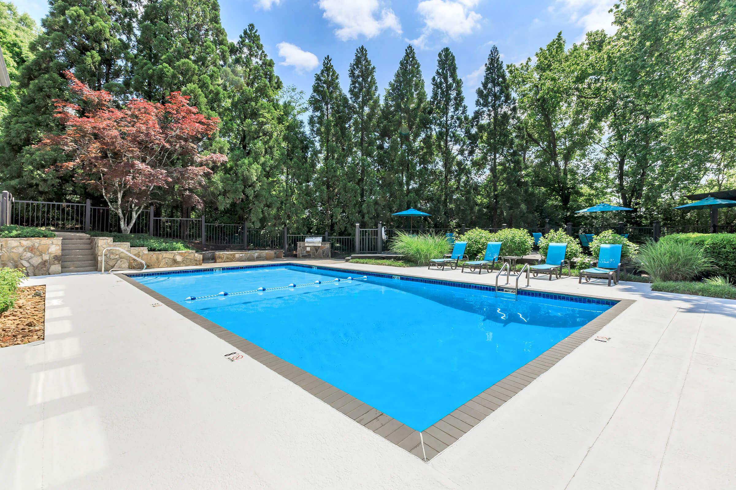 A serene outdoor pool area featuring a bright blue swimming pool surrounded by lounge chairs with turquoise cushions. Lush greenery, including tall trees and shrubs, provides privacy, while umbrellas offer shade. The setting is inviting and perfect for relaxation on a sunny day.