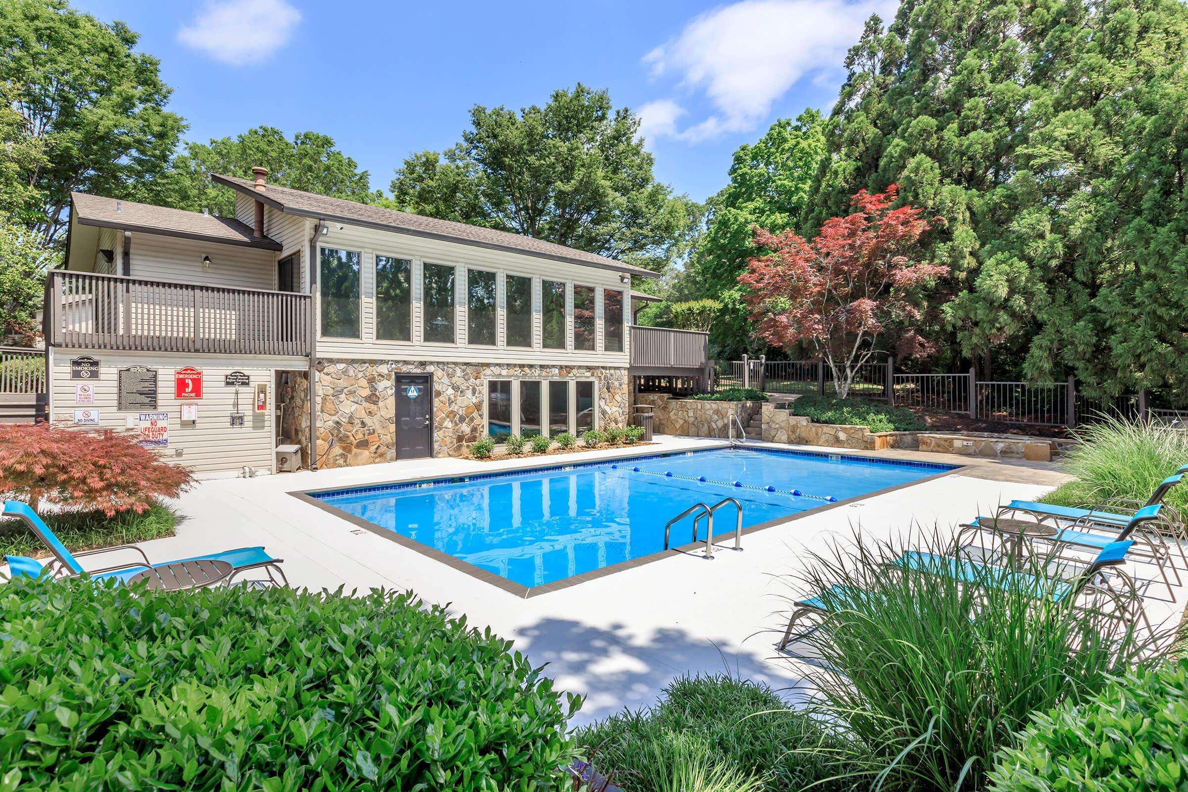 A bright outdoor swimming pool surrounded by lush greenery. The pool features blue water and a diving area, with several lounge chairs positioned around it. In the background, there is a two-story building with large windows and wooden decks. Trees and colorful shrubs provide a serene atmosphere.