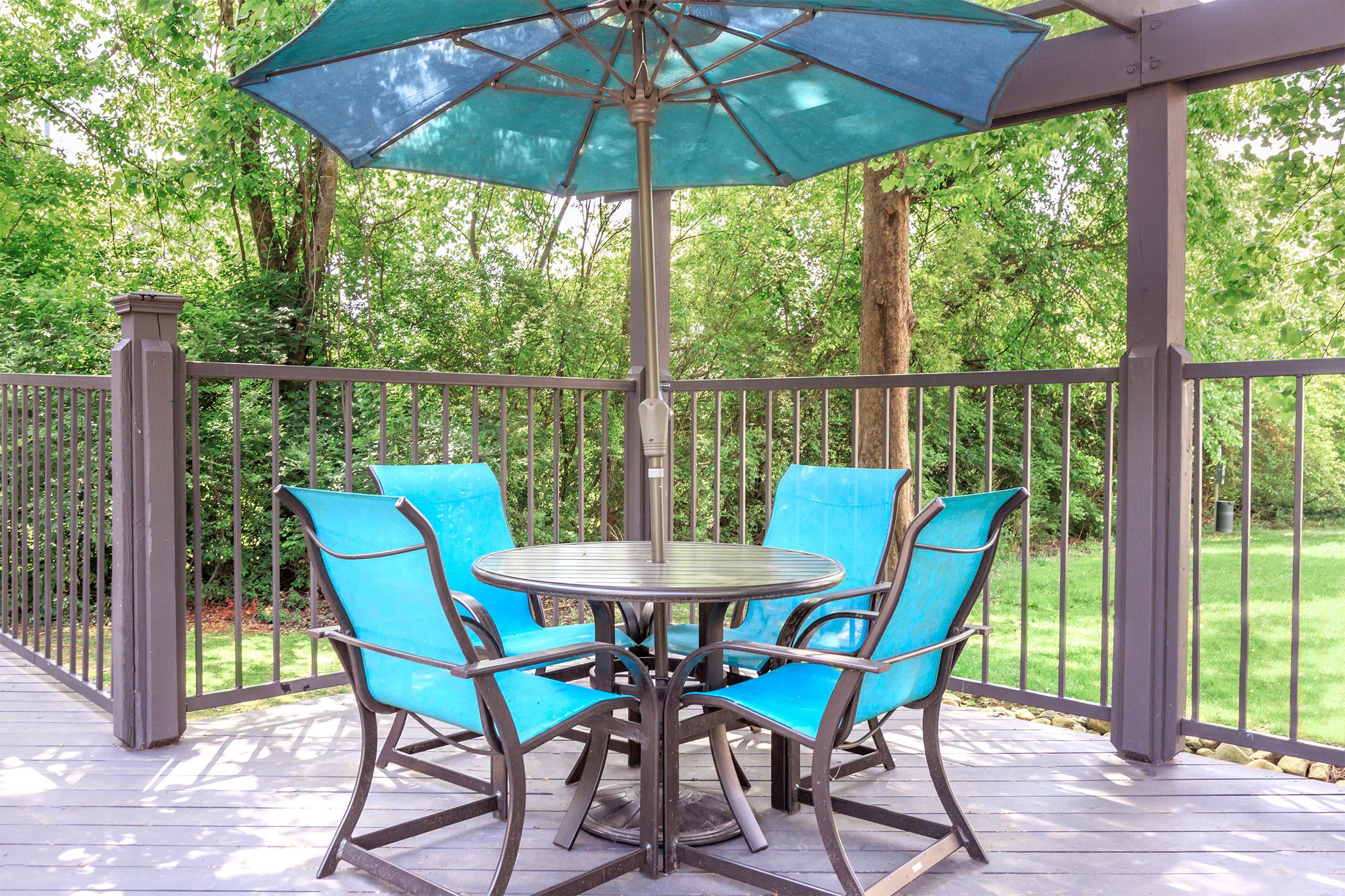 A patio set featuring a round table with a central umbrella, surrounded by four turquoise chairs, set on a wooden deck. Lush greenery in the background adds a serene outdoor atmosphere.