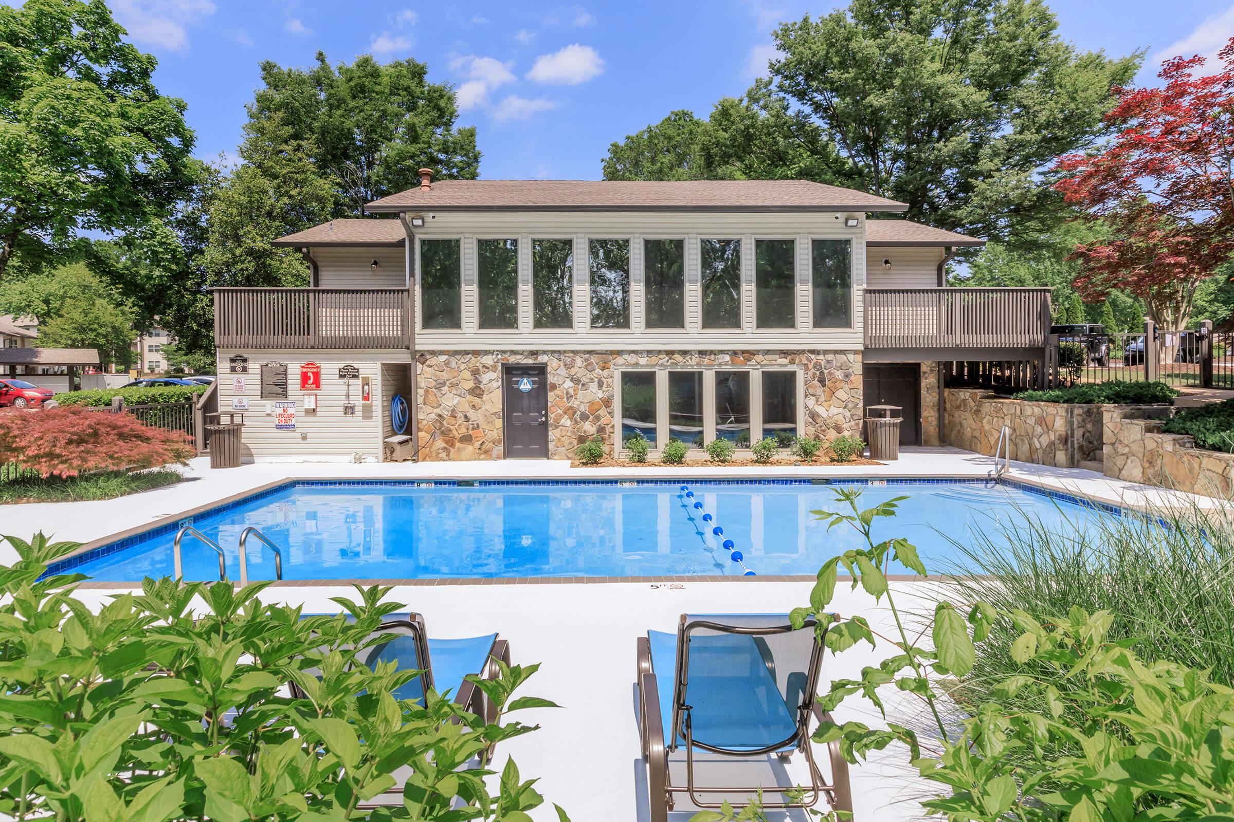 A residential swimming pool area featuring a clear blue pool surrounded by lounge chairs. In the background, a two-story building with large windows overlooks the pool. Lush green trees and colorful plants enhance the inviting outdoor space, creating a relaxing atmosphere.