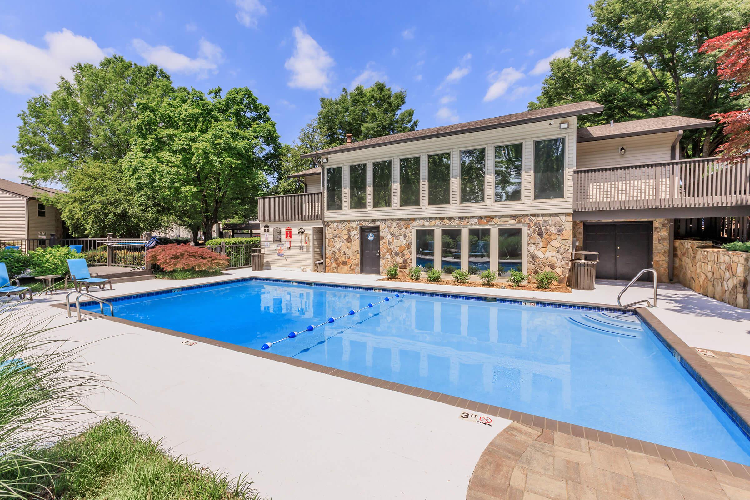 A clear swimming pool surrounded by a well-maintained landscape. In the background, there is a large building with multiple windows and a porch. Lush green trees and shrubbery enhance the outdoor setting, providing a relaxing atmosphere. The pool area features lounge chairs and safety markers.