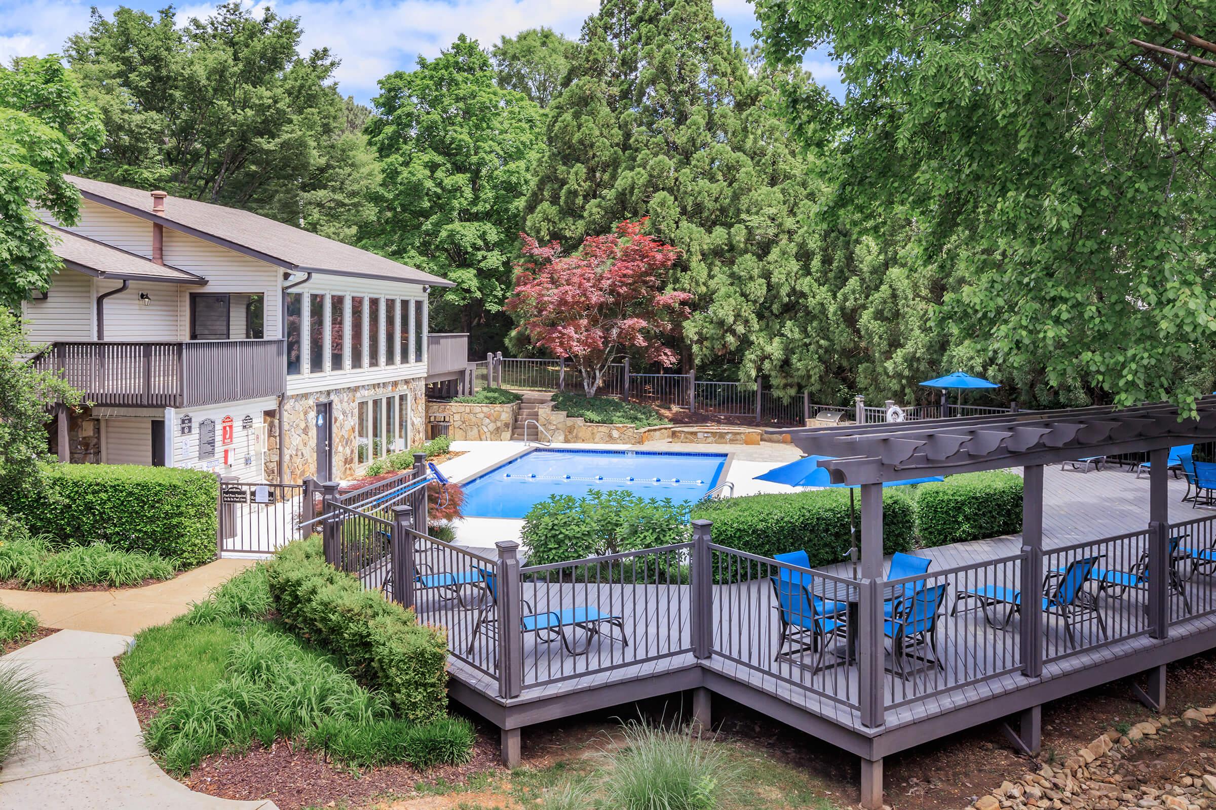 A serene outdoor pool area featuring a blue pool surrounded by wooden decking and lounge chairs. Lush greenery and trees provide shade, with a two-story building nearby that includes large windows. The scene is bright and inviting, perfect for relaxation and leisure.