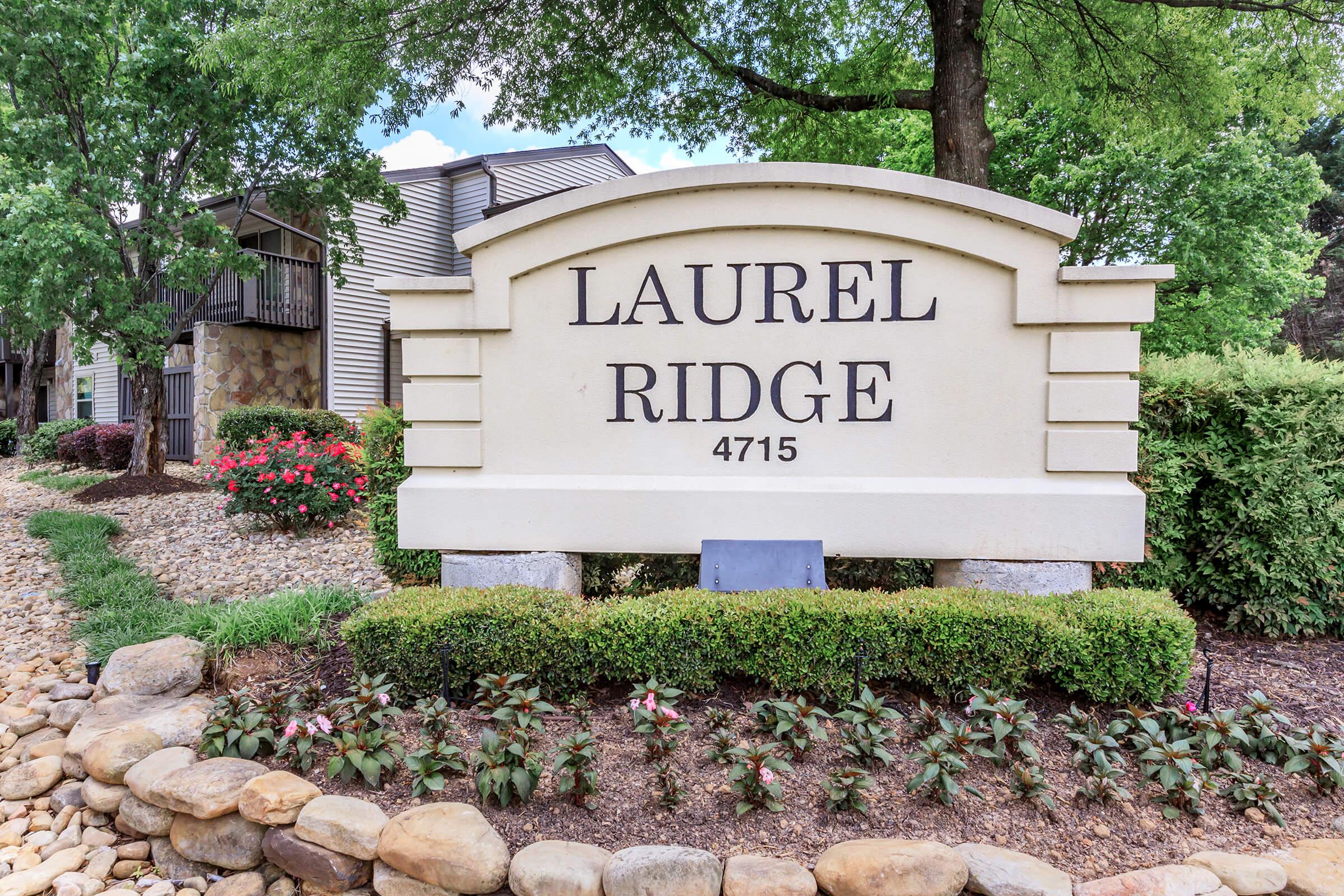 Sign for Laurel Ridge, displaying the name prominently along with the address 4715. The sign is surrounded by landscaped greenery and flowers, with a pebble pathway leading up to it. Trees provide shade in the background, creating a welcoming atmosphere.