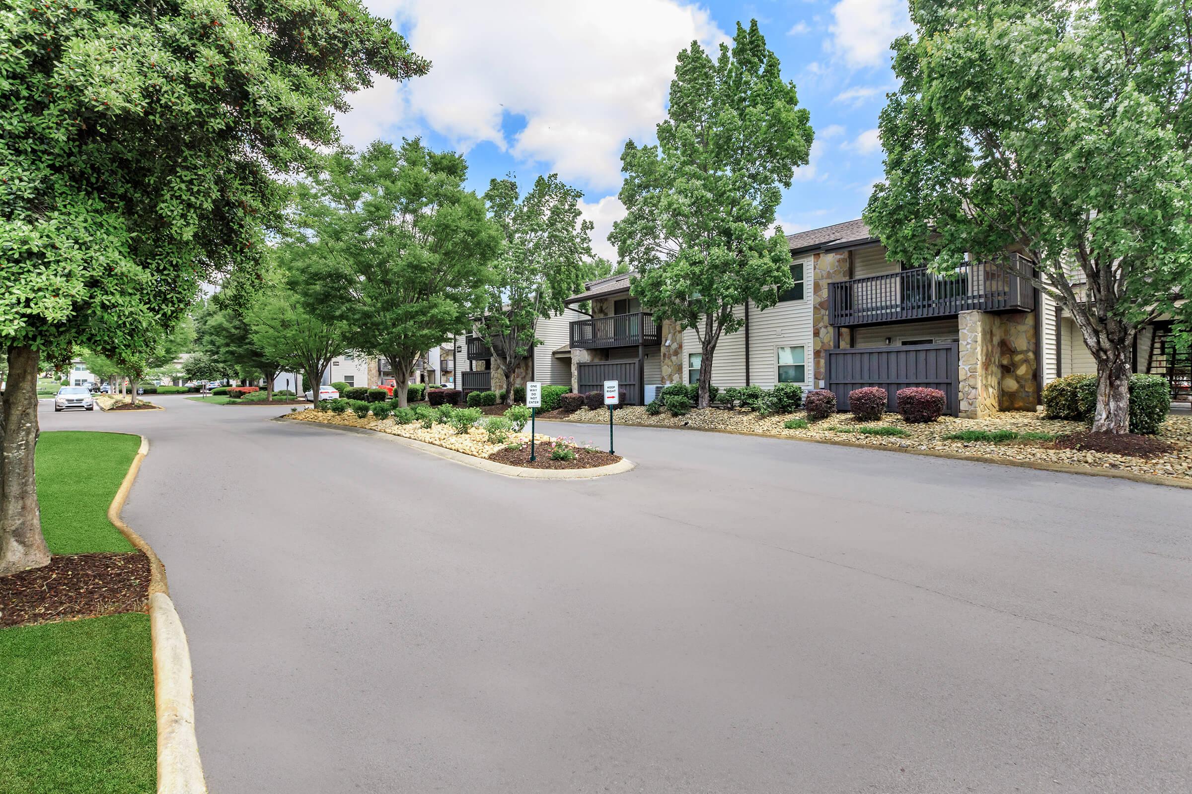 A quiet residential street lined with leafy trees and well-maintained apartment buildings. The pavement is smooth, and there are landscaped areas with shrubs and mulch. A few cars are parked along the sides, and the sky is partly cloudy, creating a peaceful atmosphere.