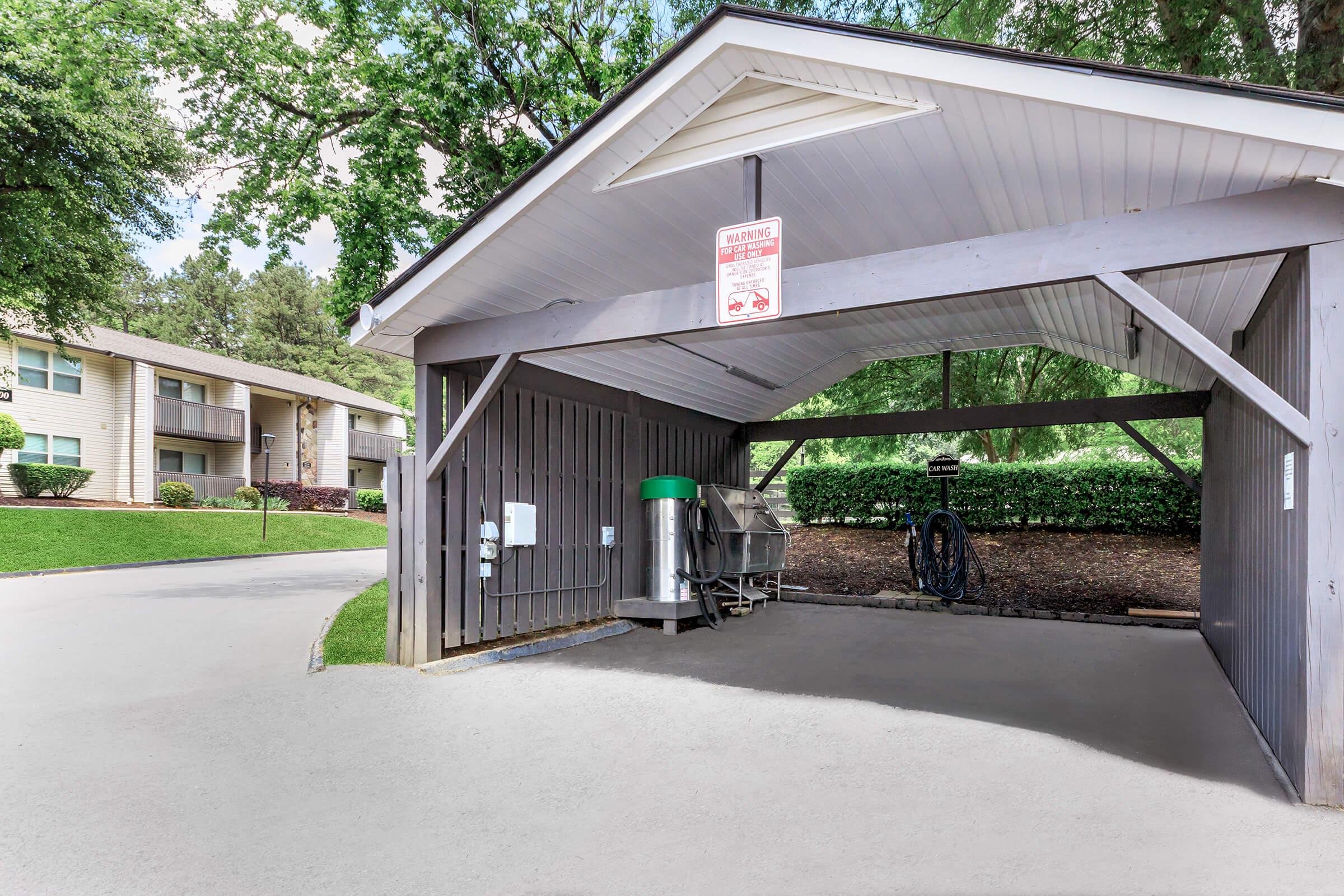 Covered bike storage area with green recycling bins and bike maintenance tools, surrounded by greenery and residential buildings in the background. The structure features wooden panels and a sign indicating storage rules. Pathway leading into the area is concrete.