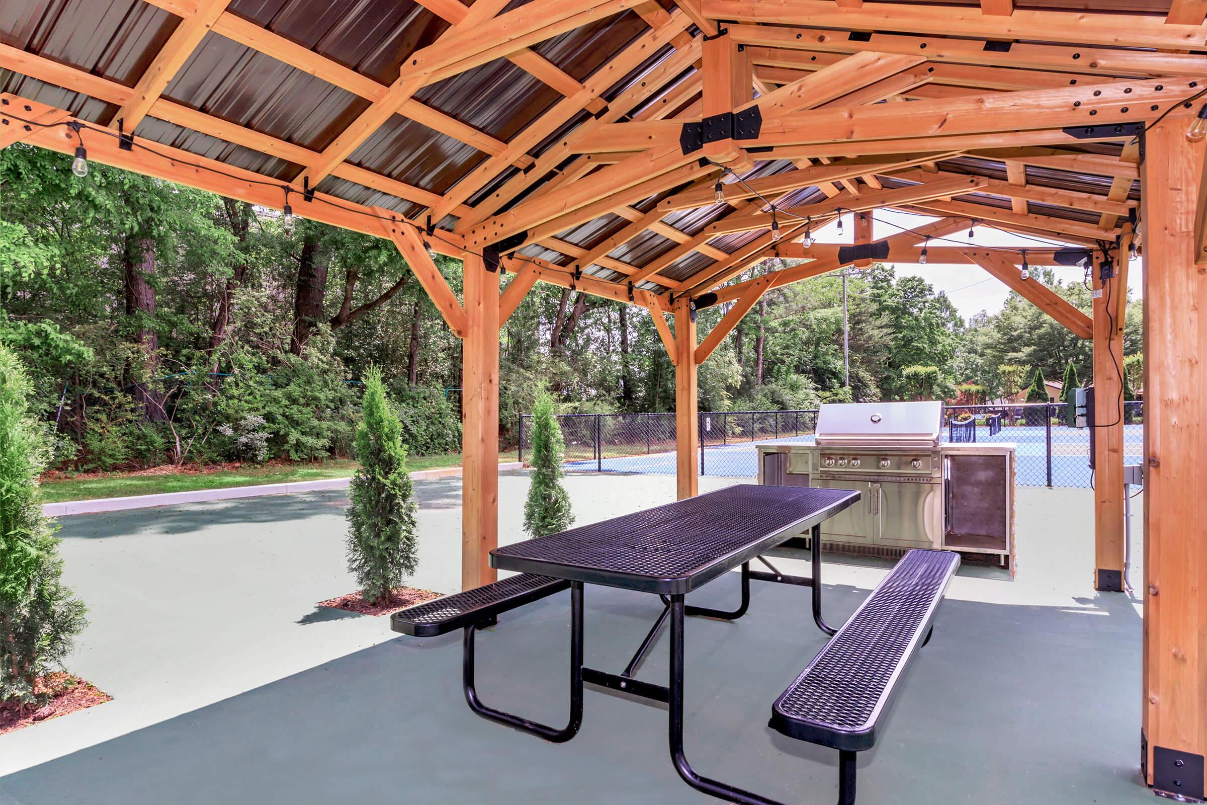 A spacious outdoor pavilion featuring a wooden structure with a metal roof. It includes a picnic table and a built-in barbecue grill. In the background, there's a tennis court surrounded by greenery, creating a relaxing recreational area.
