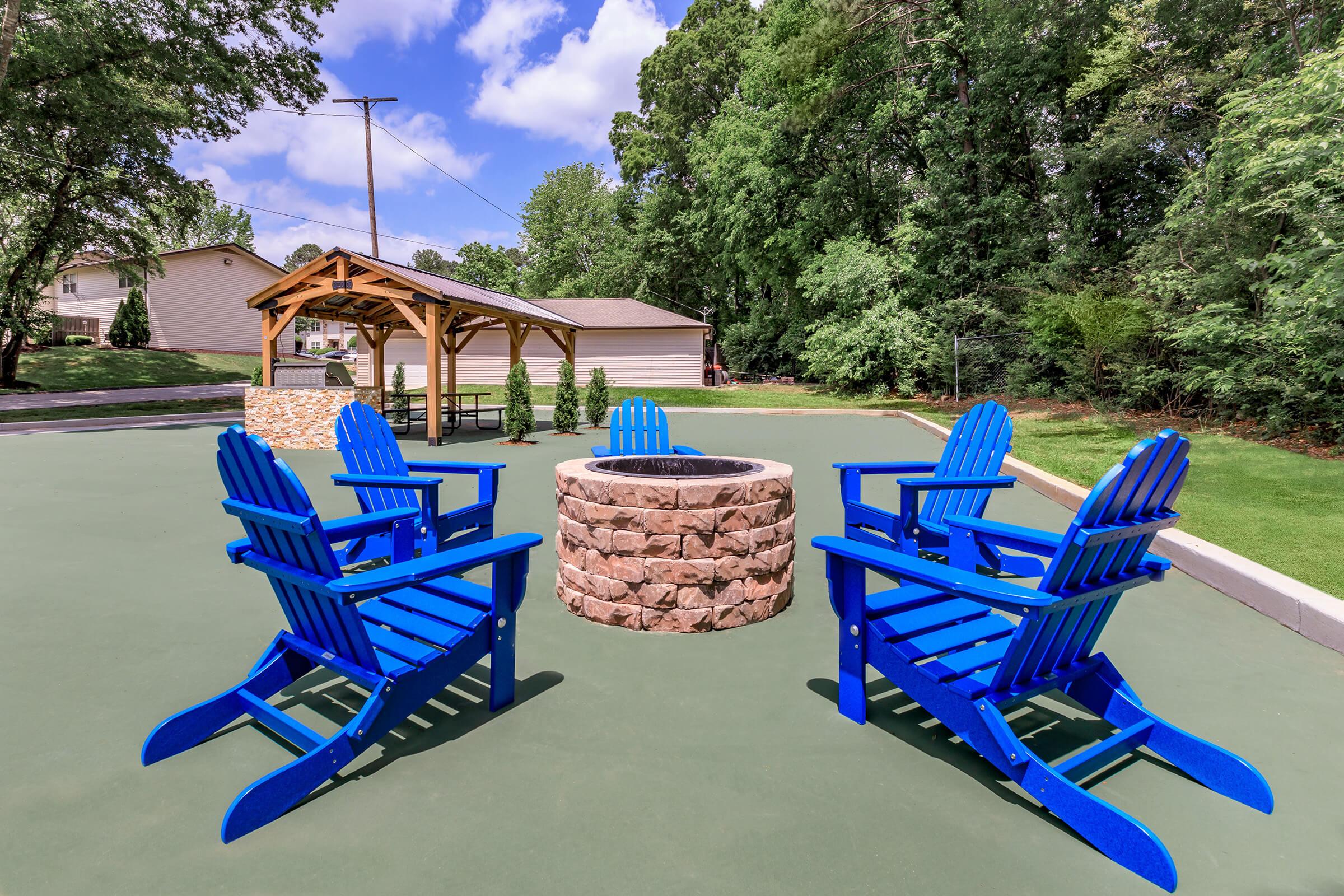 A cozy outdoor seating area featuring four bright blue adirondack chairs arranged around a circular stone fire pit. In the background, a gazebo is visible along with greenery and a blue sky, creating a welcoming space for relaxation and social gatherings.