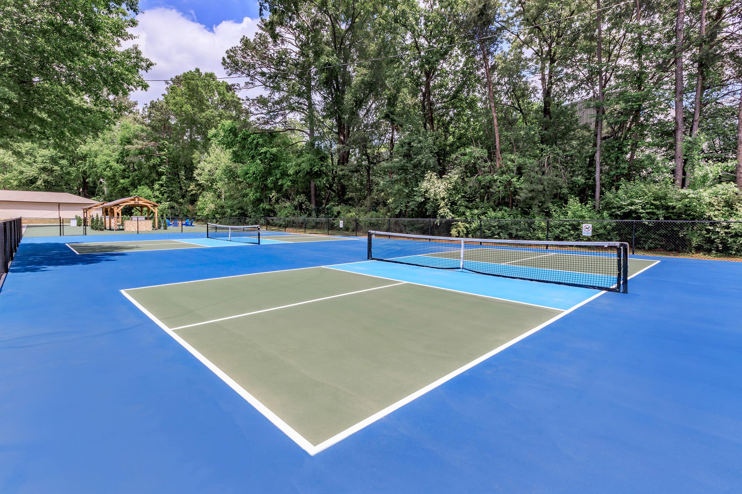 A vibrant outdoor tennis court with blue and green surfaces, surrounded by lush trees. The court features two netted areas and is well-maintained, with a clear sky above creating a bright atmosphere. Ideal for recreational play and practice.