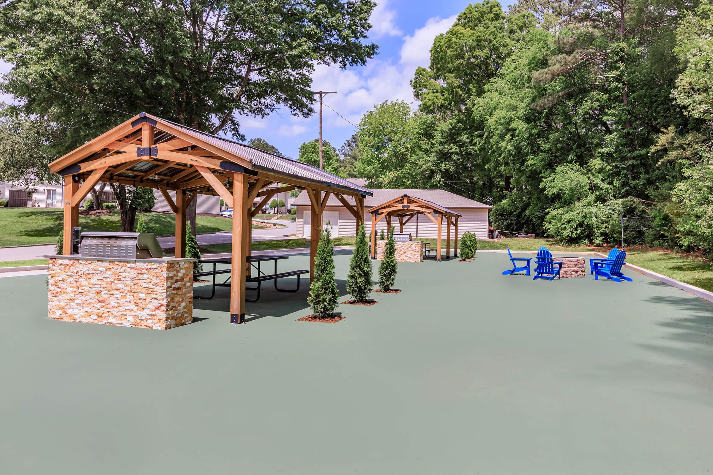 Outdoor recreational area featuring wooden gazebos with stone accents, picnic tables, and blue Adirondack chairs on a green surface. Lush trees provide shade, and the sky is partly cloudy. Ideal for gatherings and relaxation.