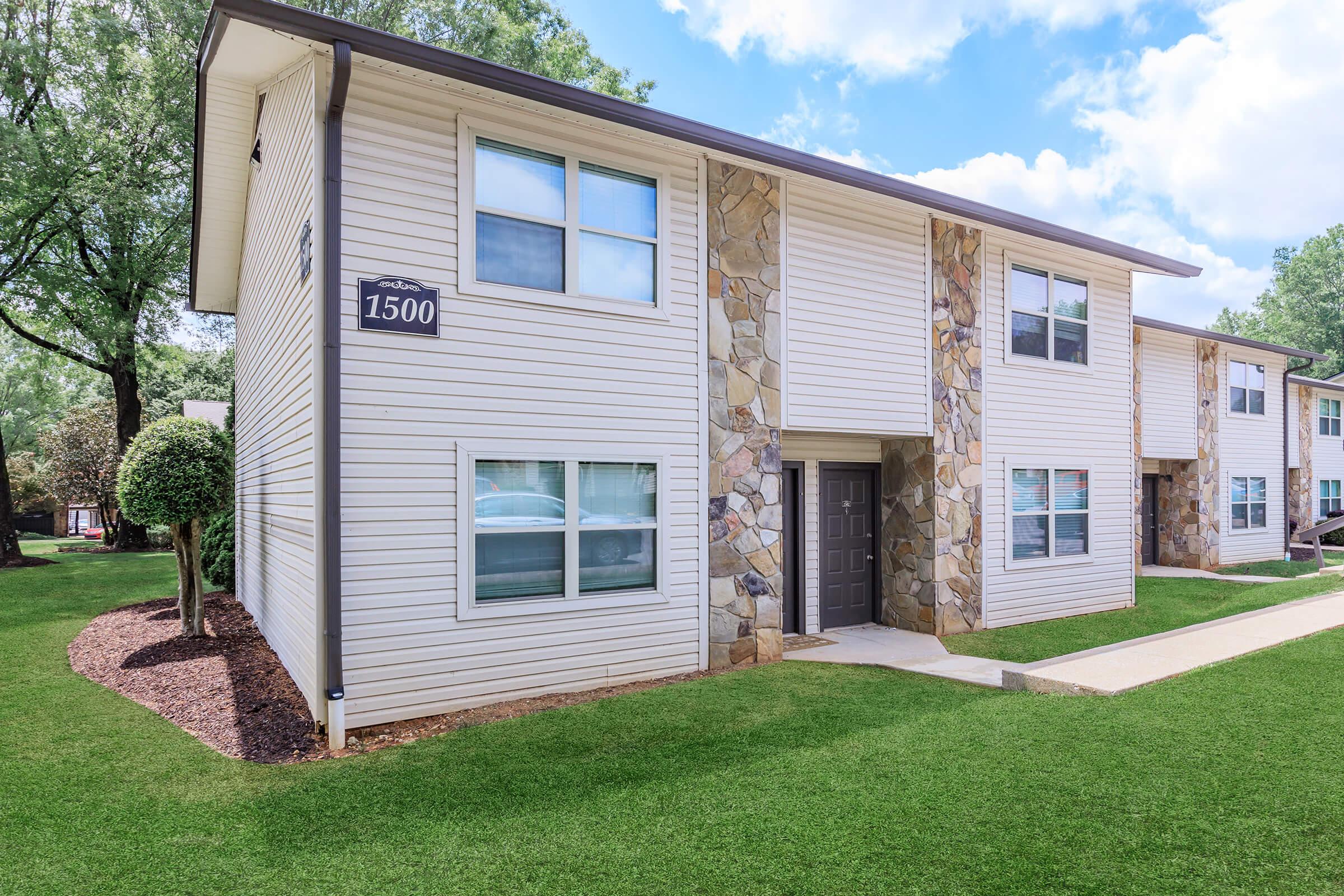 A two-story apartment building with a stone and wood exterior, featuring a numbered unit sign "1500." The front yard is landscaped with manicured grass and small shrubs, and there are cloudy skies in the background.