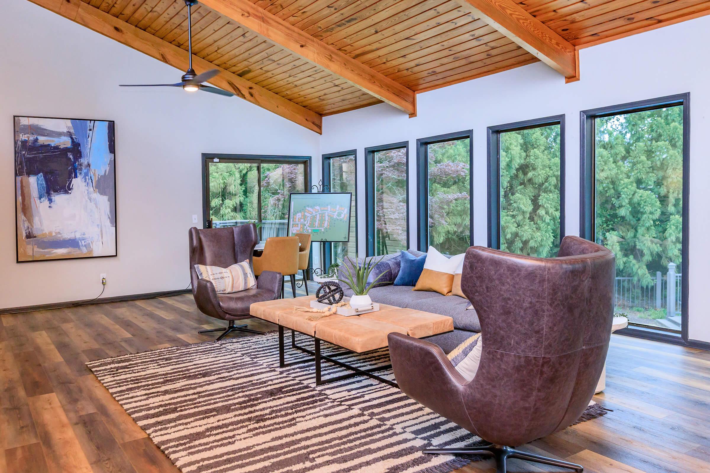 A modern living room featuring two stylish brown armchairs, a wooden coffee table with decorative items, and a large area rug with a striped pattern. Large windows provide natural light, and a wooden beam ceiling adds warmth. A piece of abstract art hangs on the wall, complementing the contemporary decor.