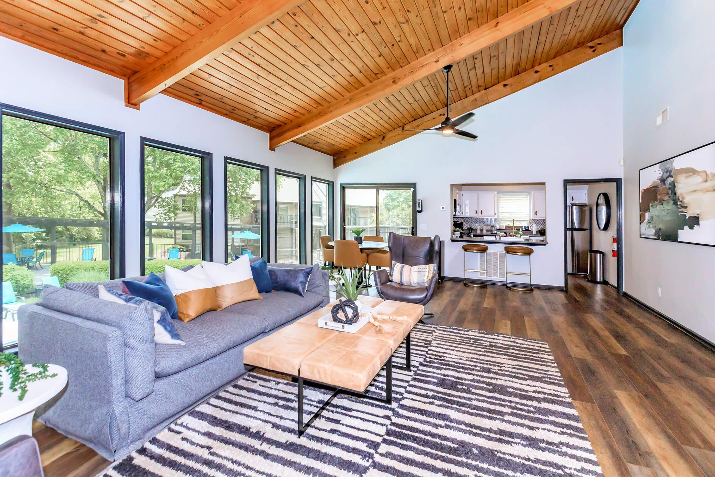 Modern living room with high wooden ceilings, large windows, and a cozy gray sofa adorned with decorative pillows. A stylish coffee table sits in the center, with a patterned rug below. In the background, a kitchen with bar stools is visible, along with outdoor seating through the windows.