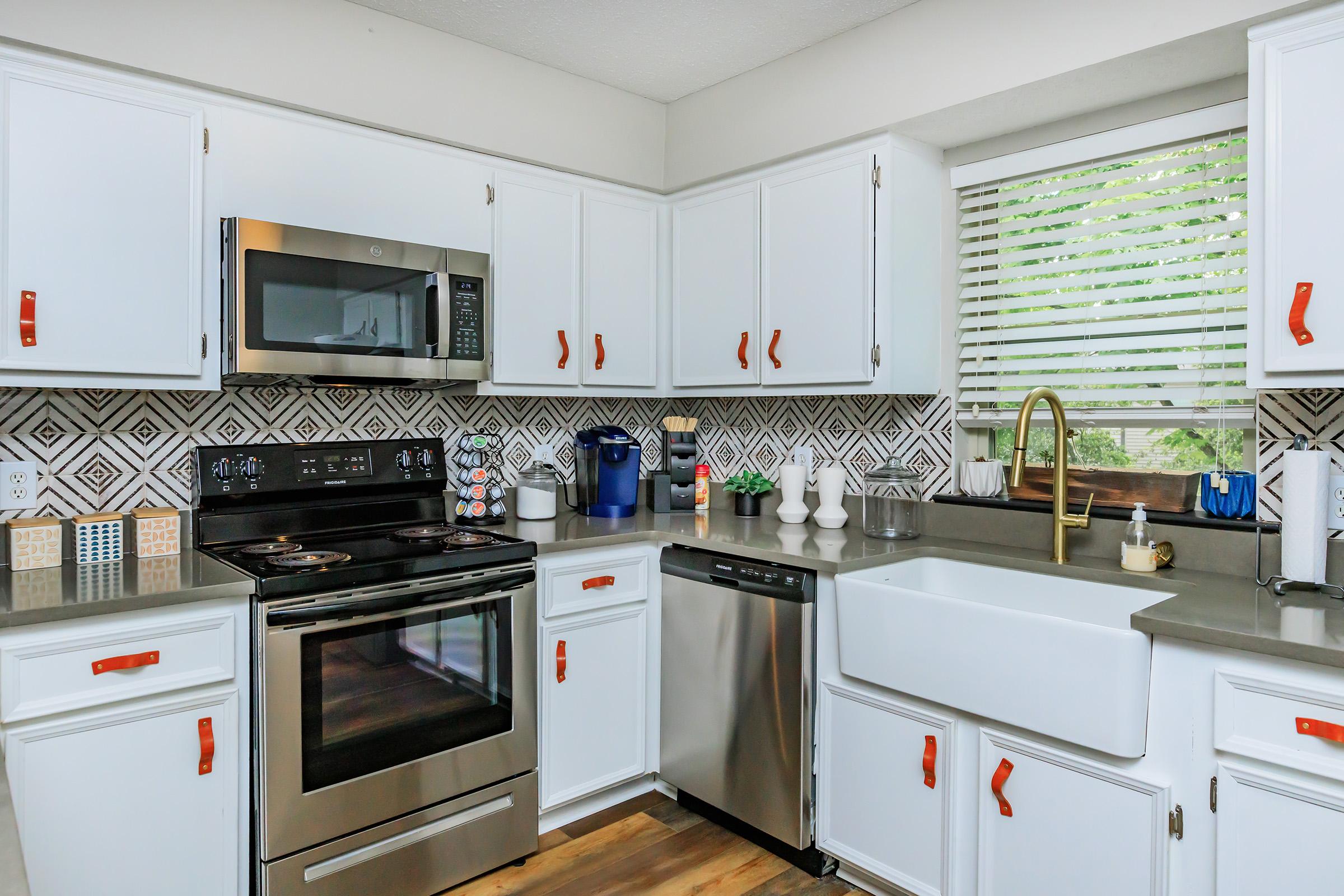 Modern kitchen featuring white cabinets with orange hardware, a stainless steel refrigerator, oven, and microwave. The countertop is gray, and there's a farmhouse sink below a window with blinds. Decorative items are displayed, including a coffee maker and containers on the counter.