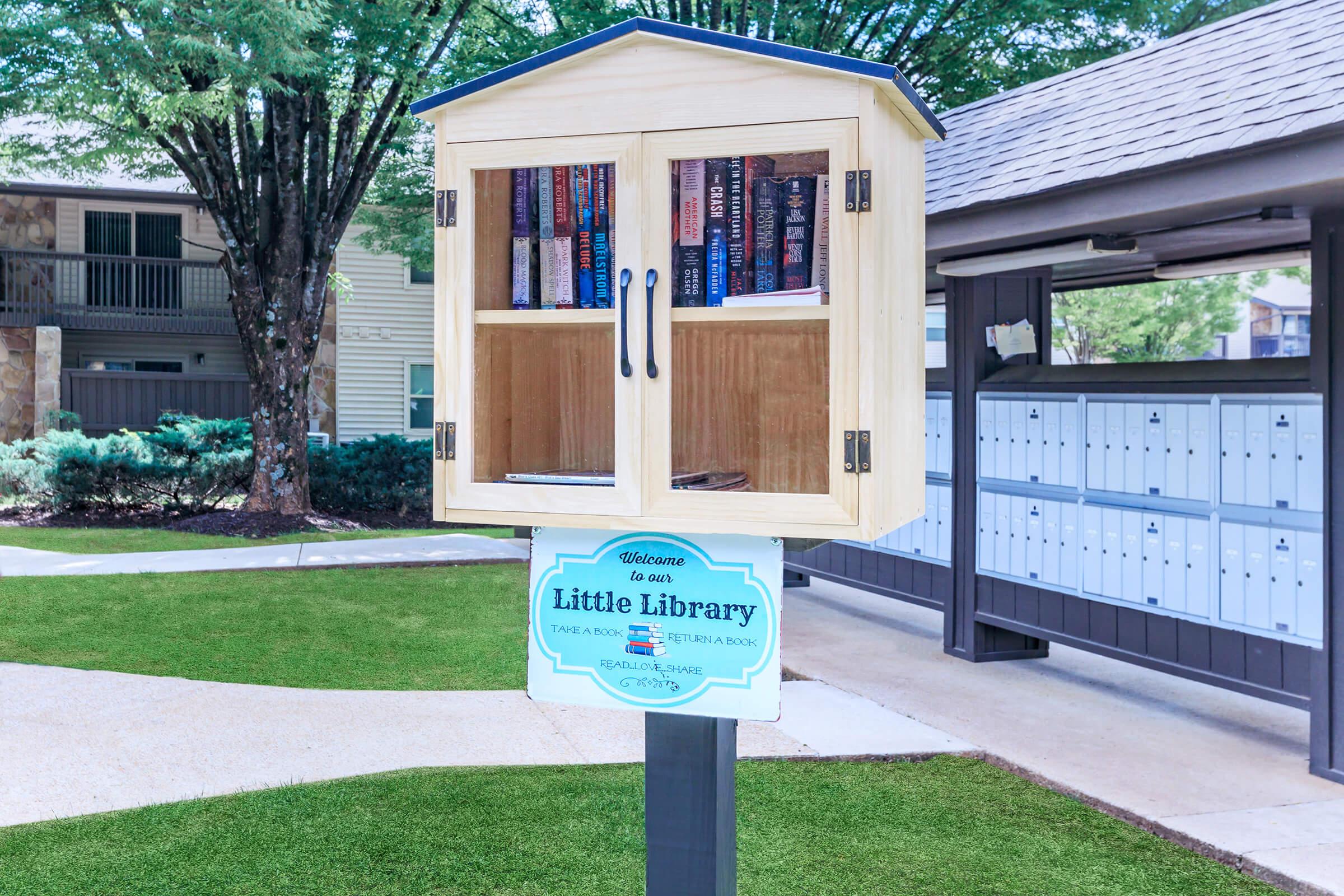 A wooden little library box filled with books, placed on a post in a grassy area. The library has glass doors and a sign that reads "Welcome to our Little Library" with a message about borrowing and donating books. Surrounding trees and a mail area are visible in the background.