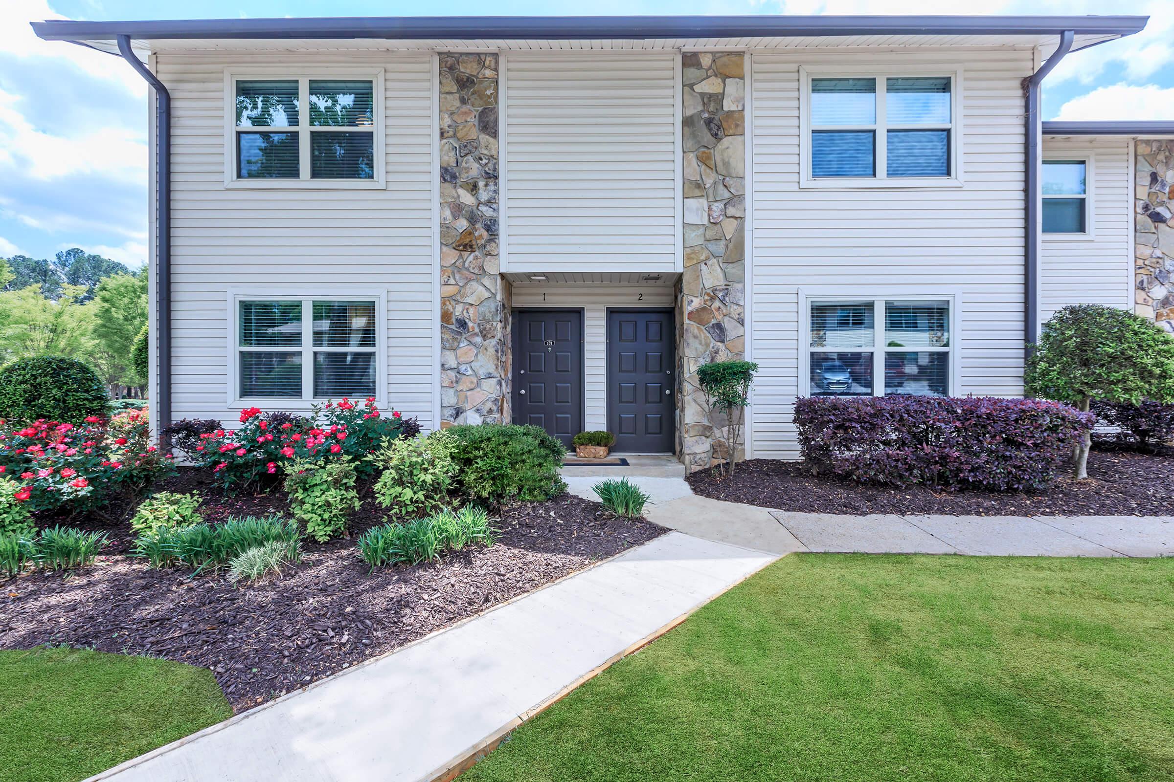 A well-maintained apartment building with a stone and siding exterior. The front features two dark doors framed by decorative plants and colorful flower beds. A neatly trimmed lawn and paved walkway lead to the entrance, with blue skies and fluffy clouds in the background.