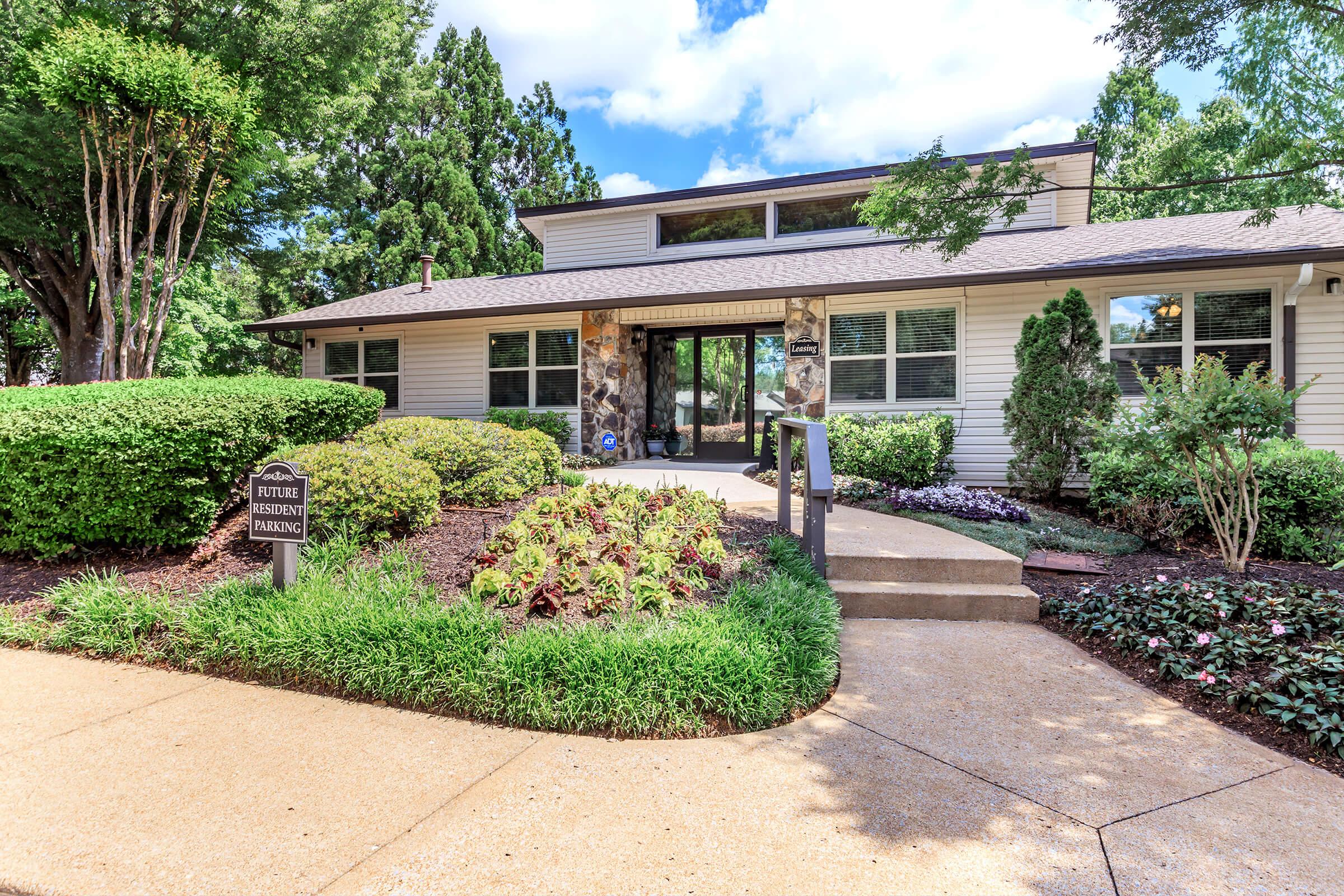 A modern building surrounded by well-maintained landscaping, featuring a stone pathway and a sign that reads "Future Resident Parking." Lush green bushes and colorful plants enhance the entrance, with a clear blue sky overhead and trees in the background.