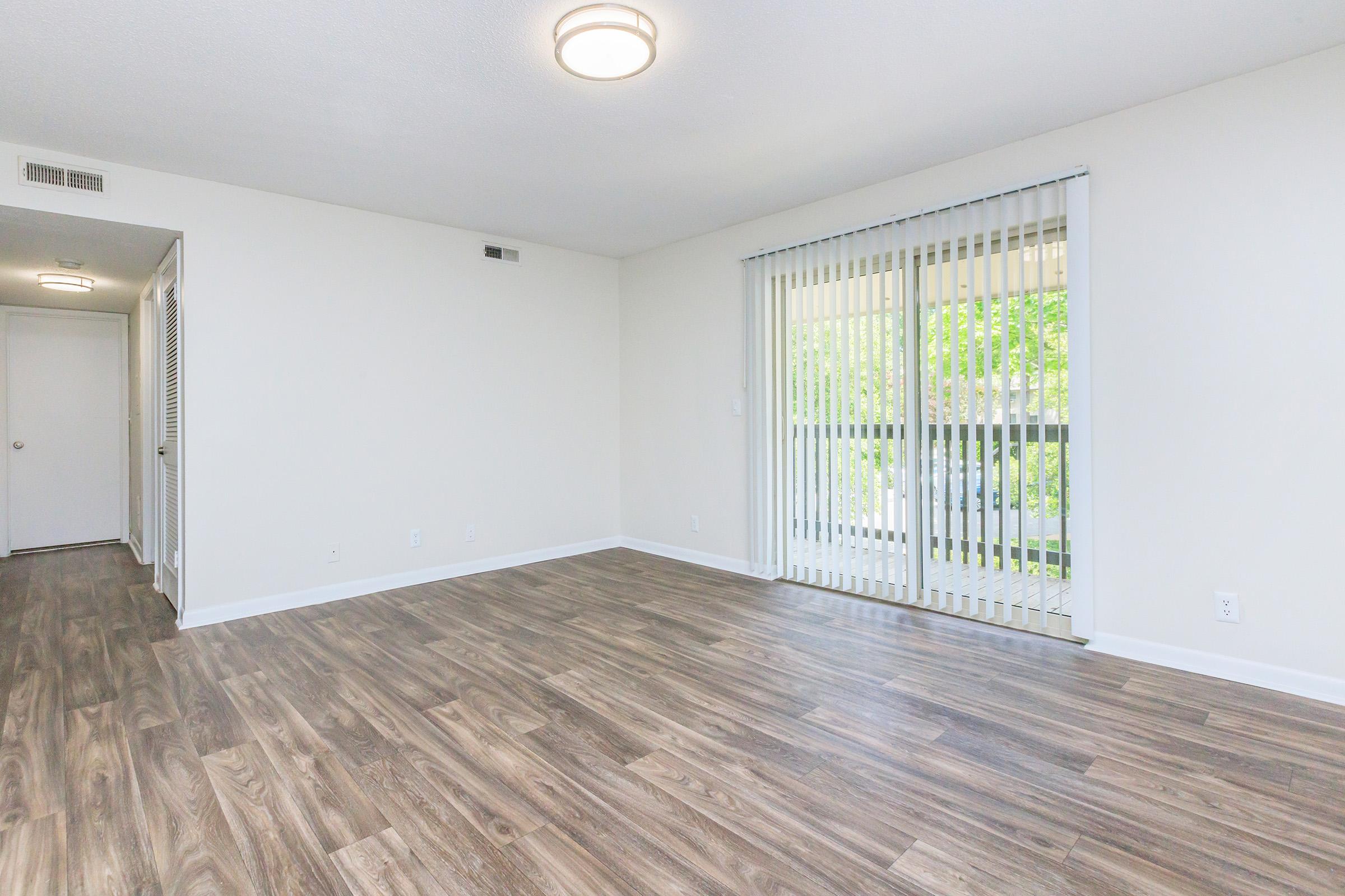 Spacious and empty living area featuring light-colored walls, hardwood-style flooring, and a large sliding glass door with vertical blinds that opens to a balcony or outdoor space. A door leads to a hallway, and there is ample natural light illuminating the room.