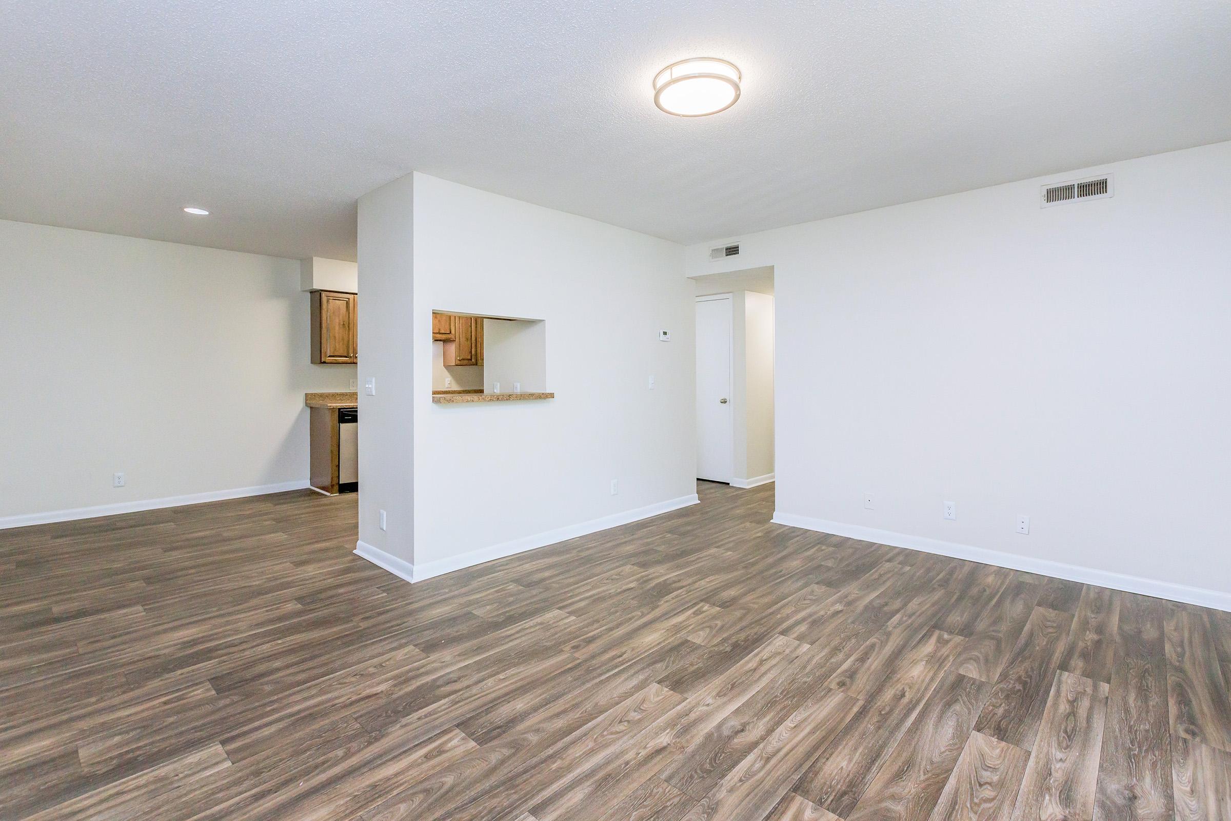 Spacious living area featuring light wood-style flooring, neutral-colored walls, and modern lighting. A partial wall with a pass-through connects to a kitchen area partially visible in the background. The room is bright and empty, suitable for personalization.