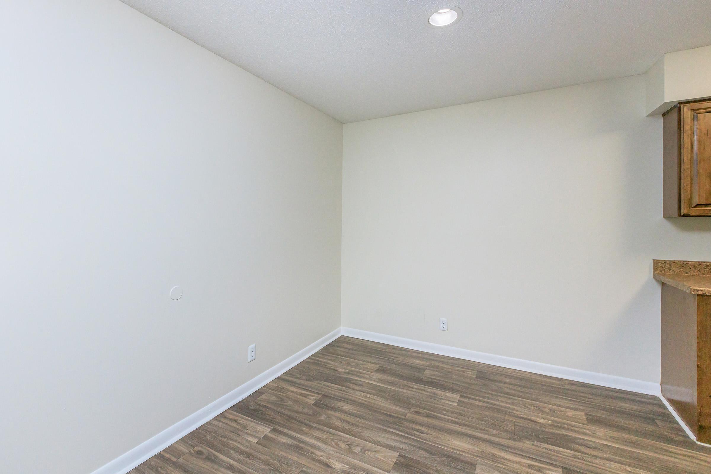 A small, empty room with light-colored walls and a wood-style floor. There is a small portion of a kitchen visible to the right, featuring wooden cabinets and a countertop. The lighting is warm and there is a recessed ceiling light.